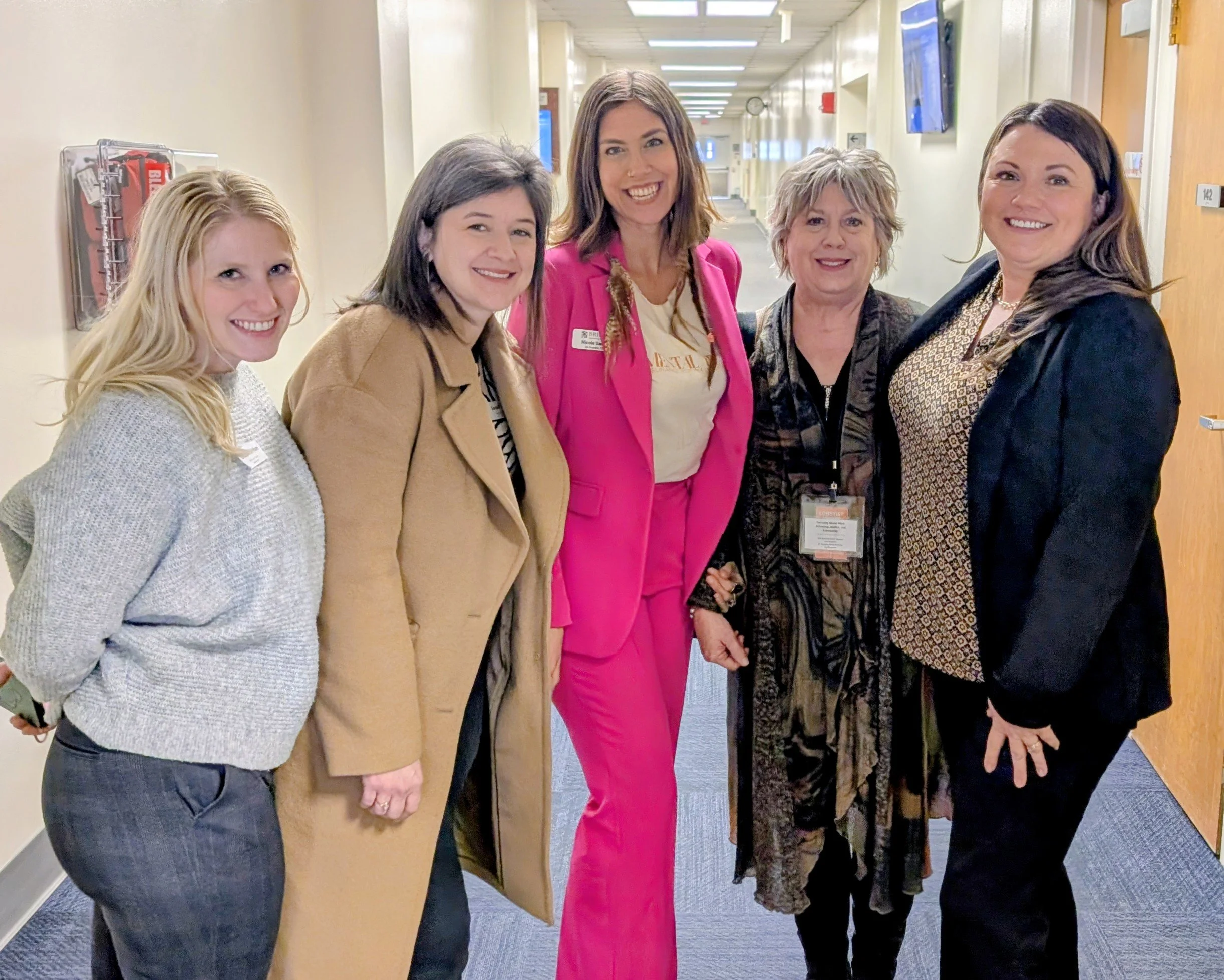 Five women standing together and smiling in a hallway