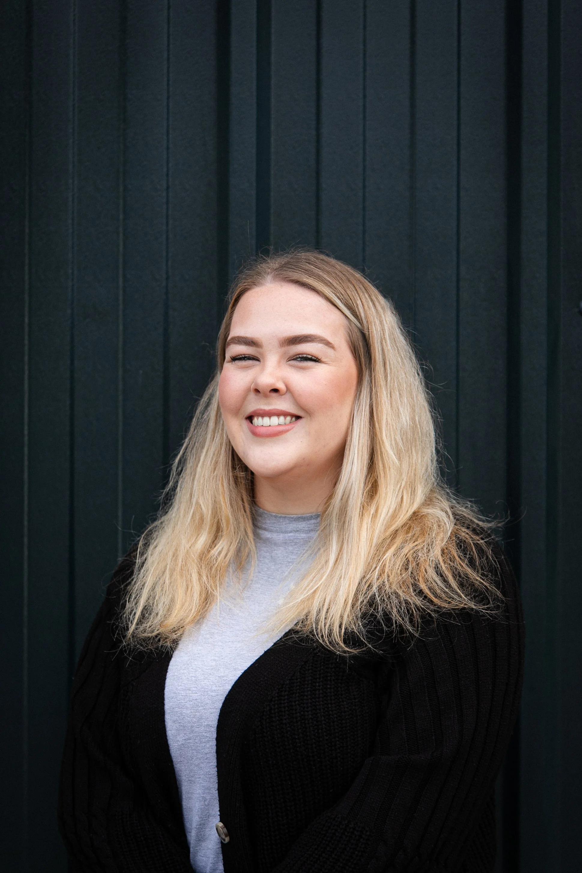 A young woman with long blonde hair, smiling and winking, standing in front of a dark green textured wall.
