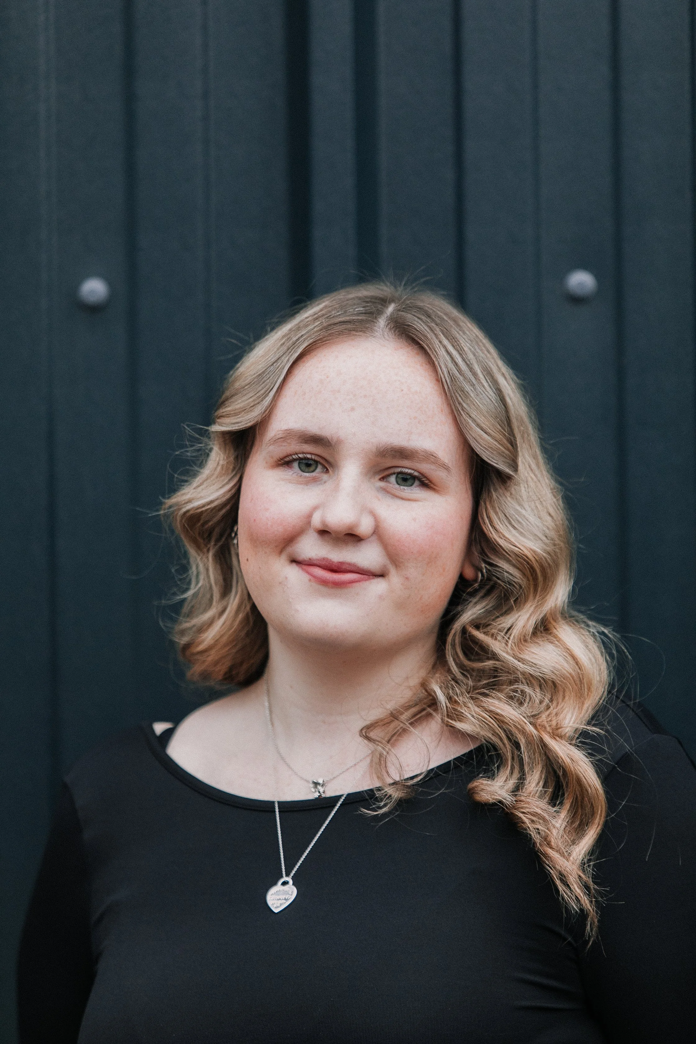 A young woman with wavy blonde hair, wearing a black top and silver necklaces, standing in front of a dark textured background.