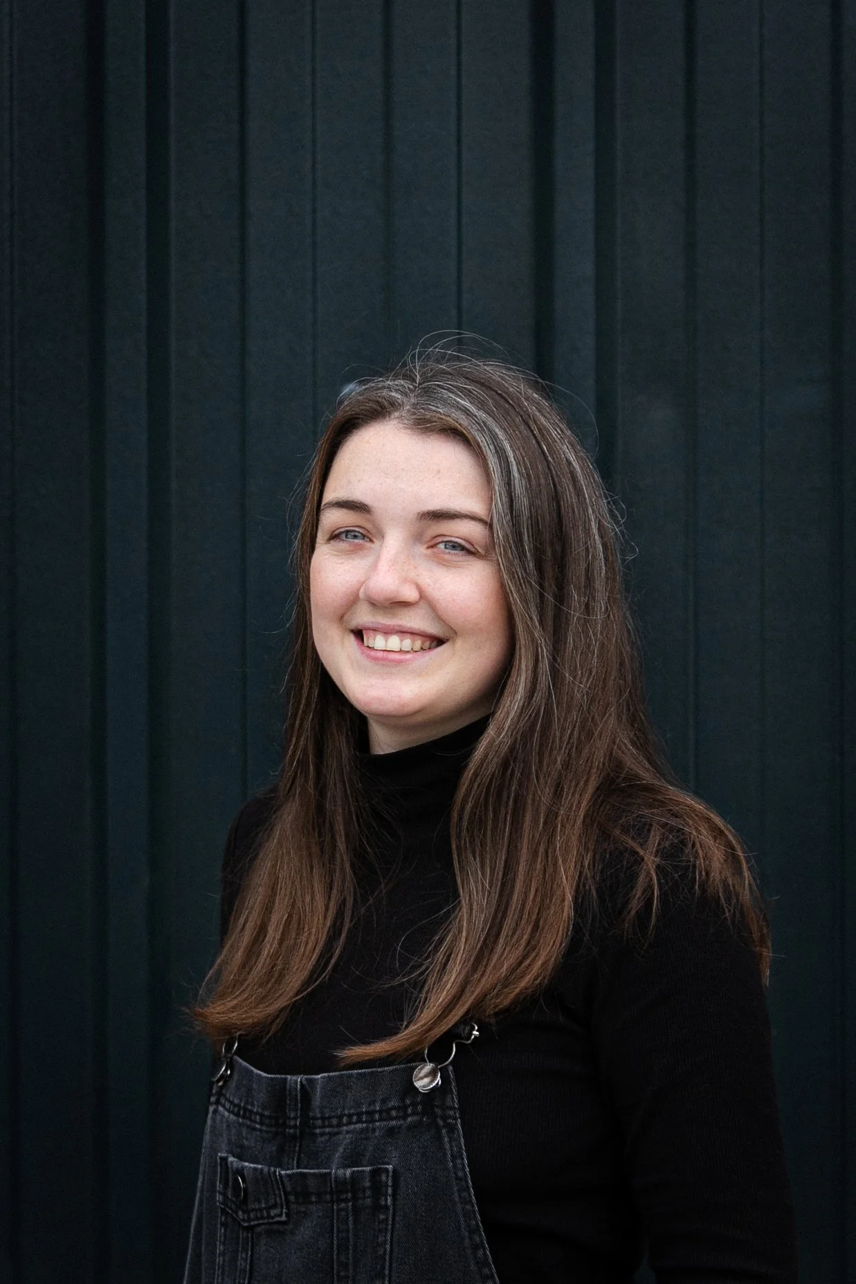 A young woman with long brown hair smiling, wearing a black turtleneck and black overall, standing in front of a dark green wooden wall.