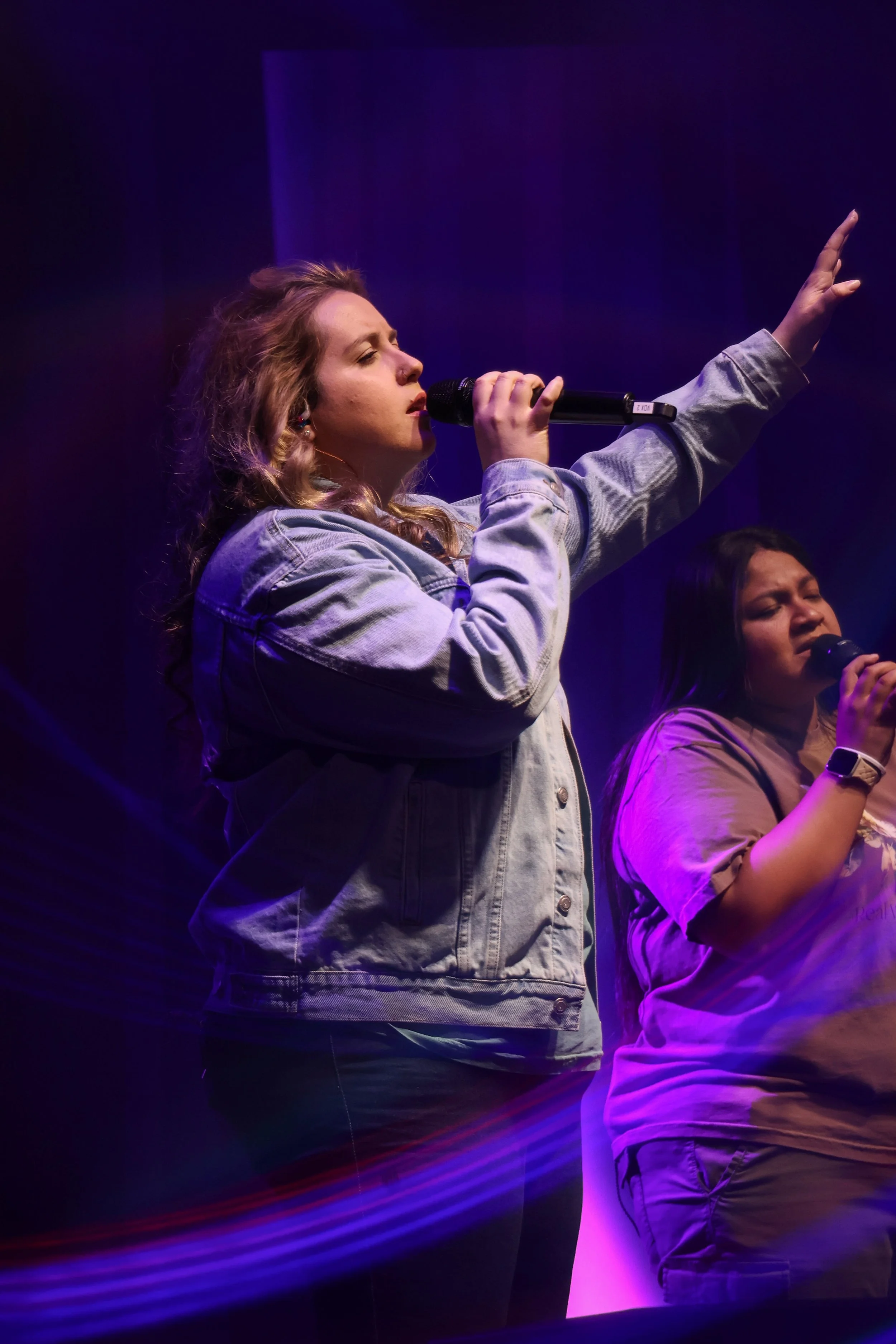 Two women singing on stage with microphones, one wearing a denim jacket and the other a beige shirt, illuminated by purple and pink stage lights.