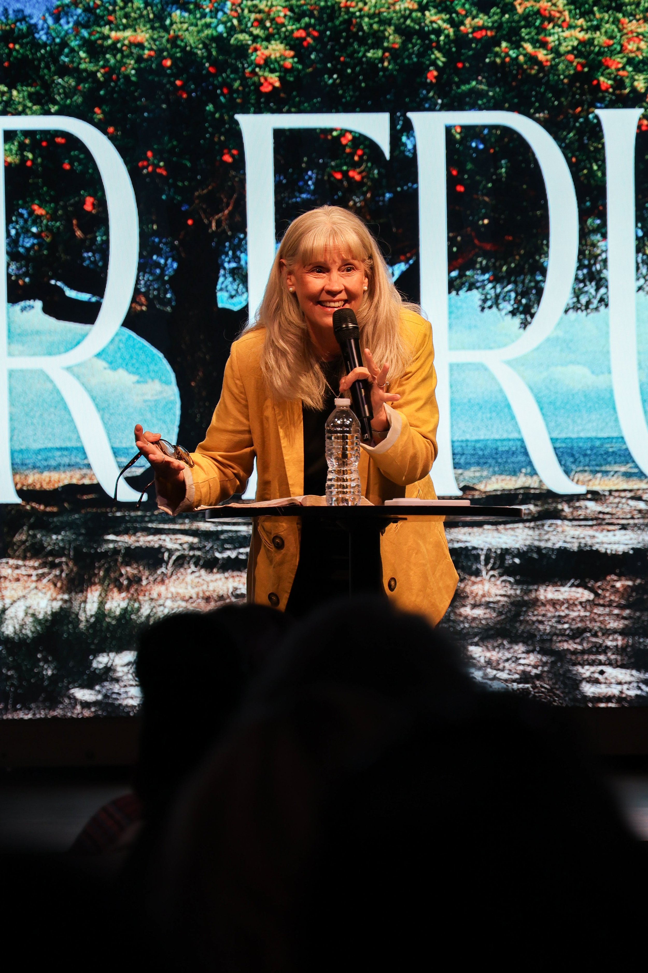A woman with blonde hair wearing a yellow jacket is speaking into a microphone during a presentation or talk, standing at a small table with a bottle of water and glasses, against a large backdrop featuring an image of a large tree and blue sky.
