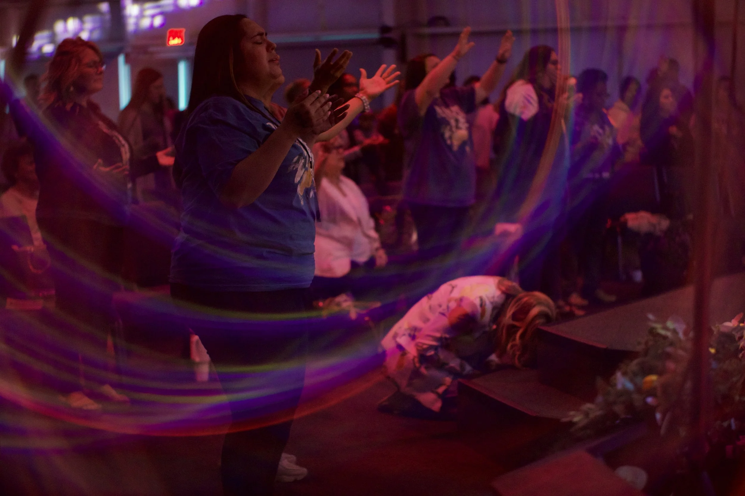 People praying and worshiping in a church, with some standing with eyes closed and hands raised, and a woman kneeling at the altar.