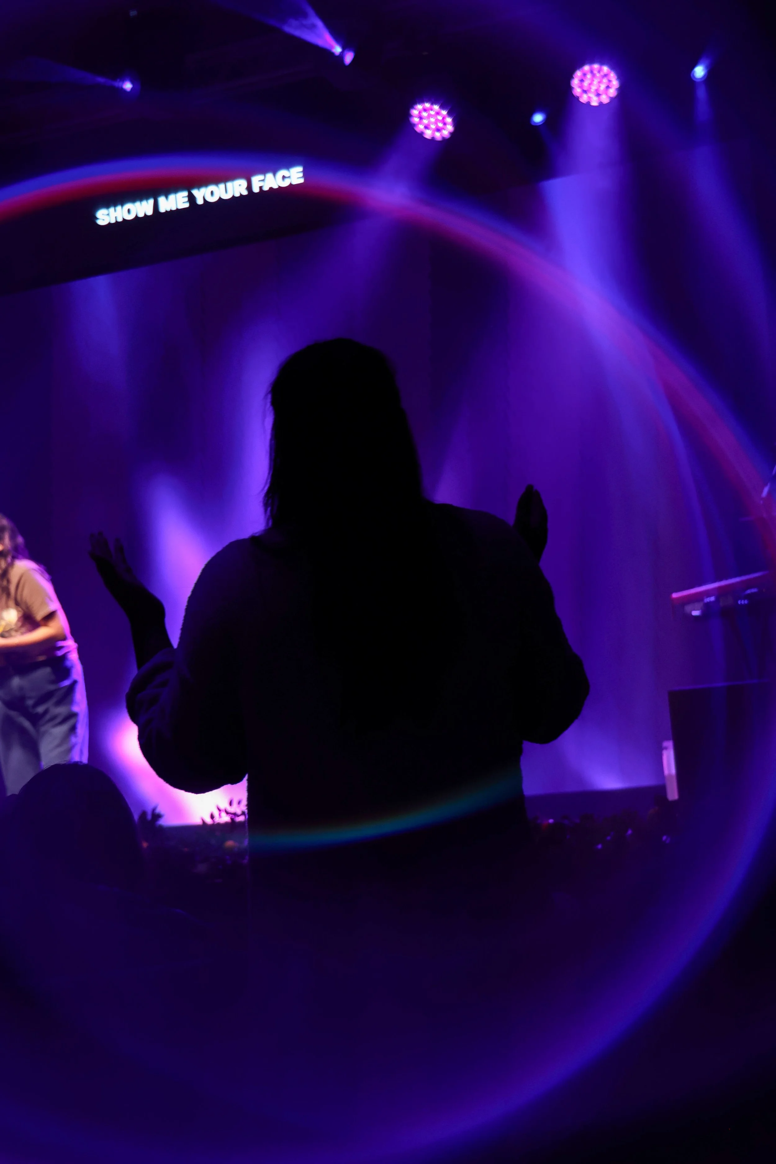 A person dancing in a dark room with purple and blue stage lights, and a screen displaying the phrase 'SHOW ME YOUR FACE'.