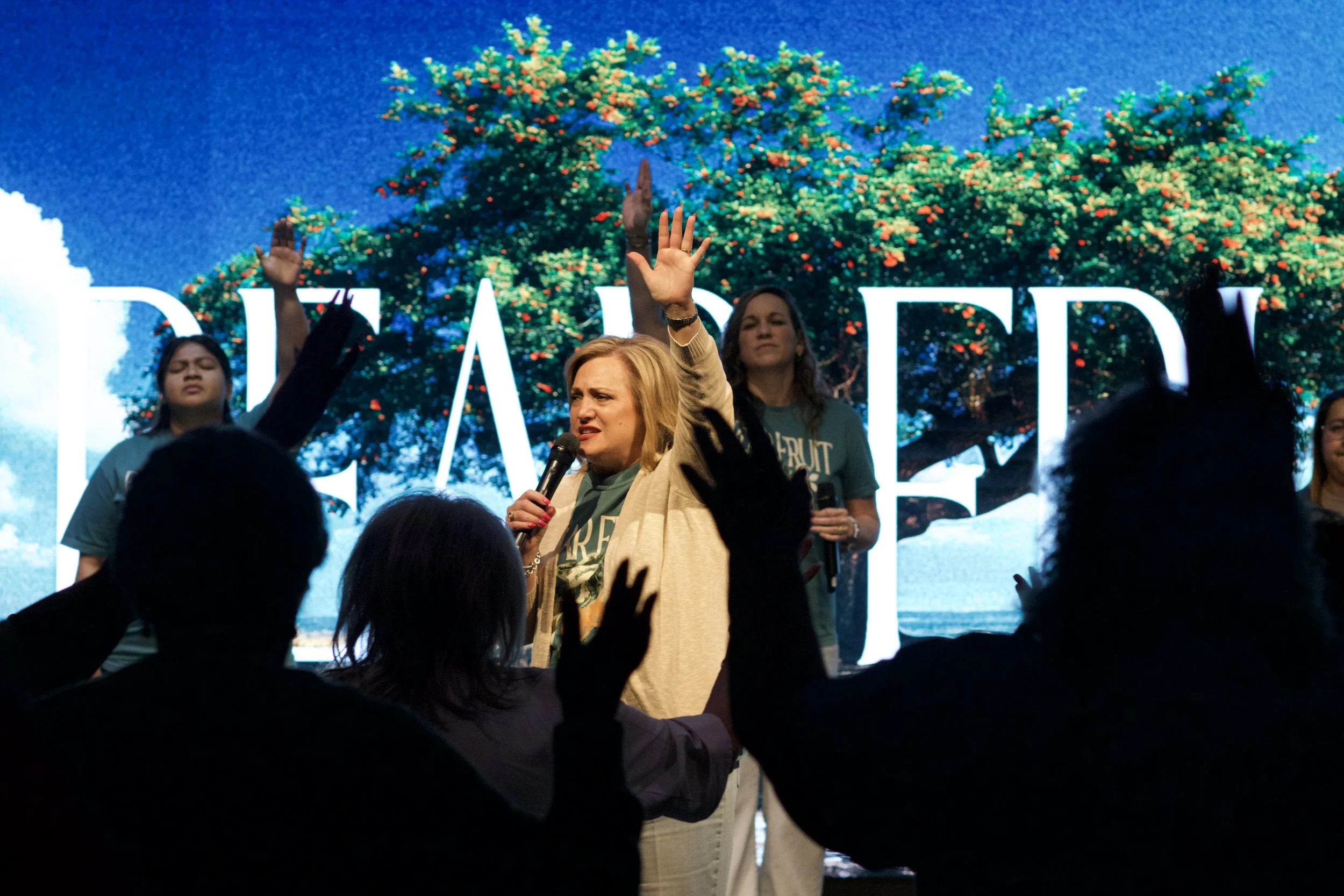 A woman with blonde hair speaking into a microphone with her right hand raised during a presentation or event, with a background displaying large text and an image of a large tree.