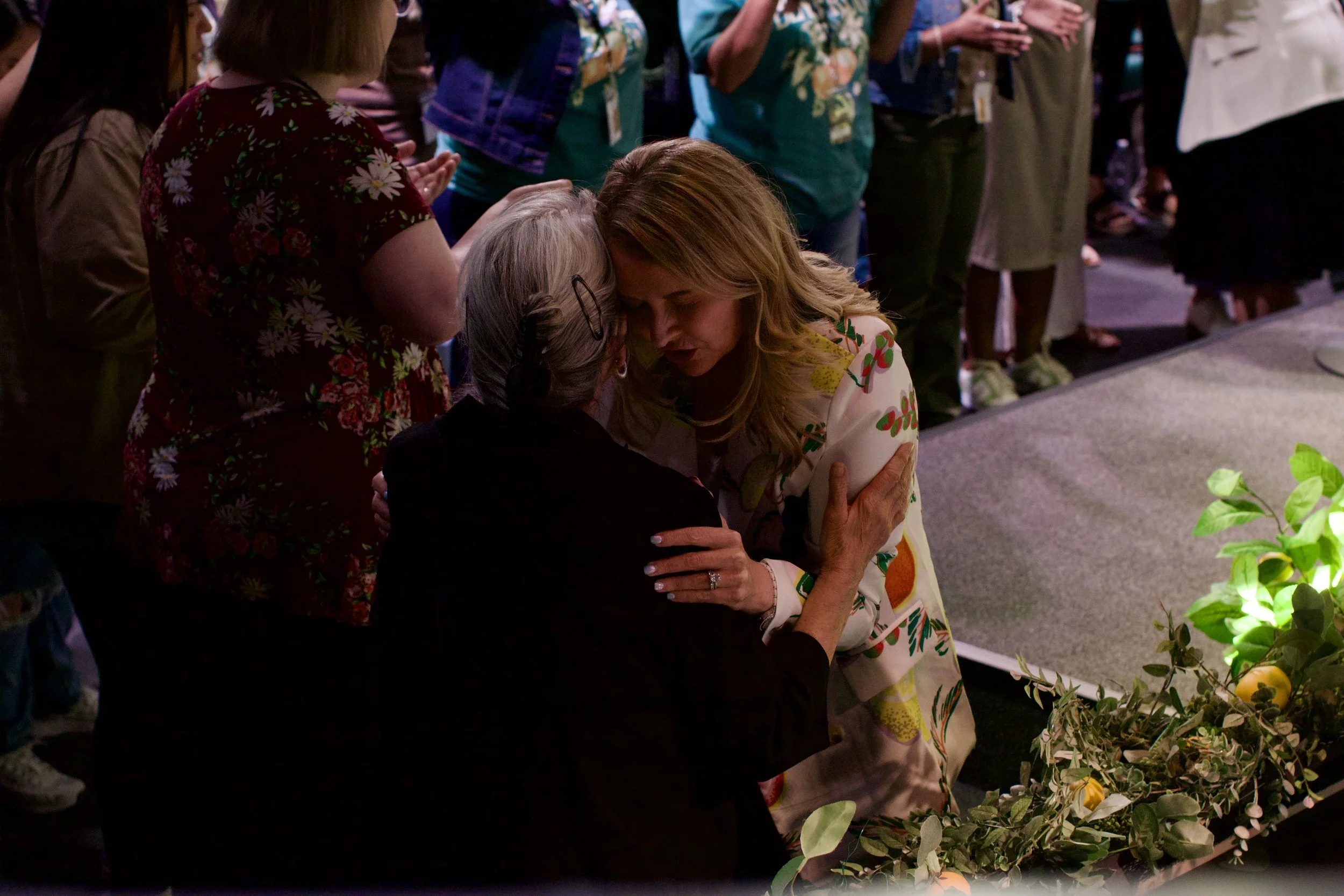 A woman with long red hair wearing a white dress with colorful fruit patterns embraces an elderly woman with gray hair and glasses at a gathering, with several people standing nearby.