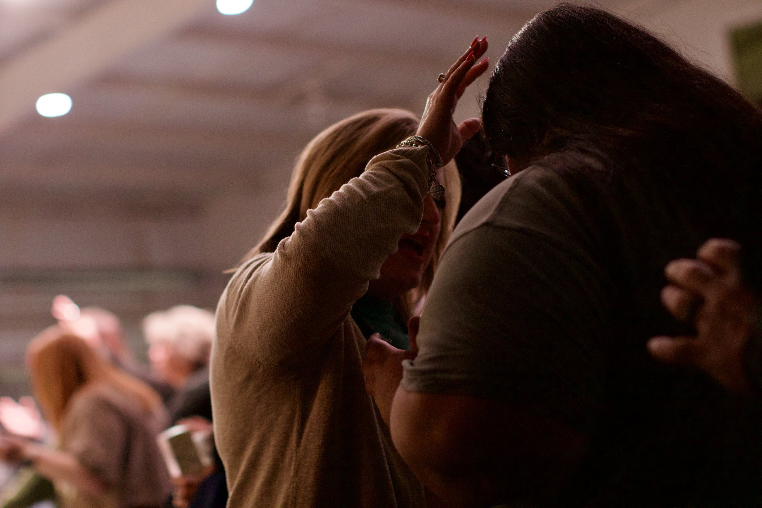 Two women in an emotional moment, touching foreheads in a crowded indoor setting.