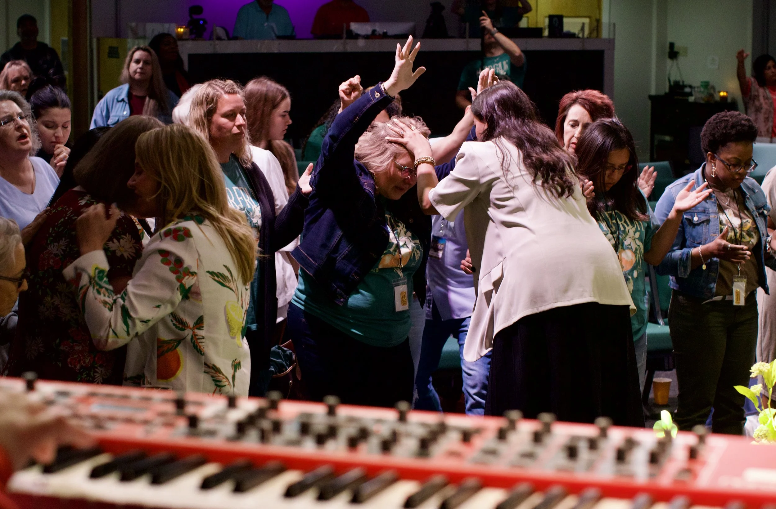 A group of diverse women standing in a circle with their eyes closed and hands raised, engaged in prayer or worship in a room with music equipment and a blurred keyboard in the foreground.