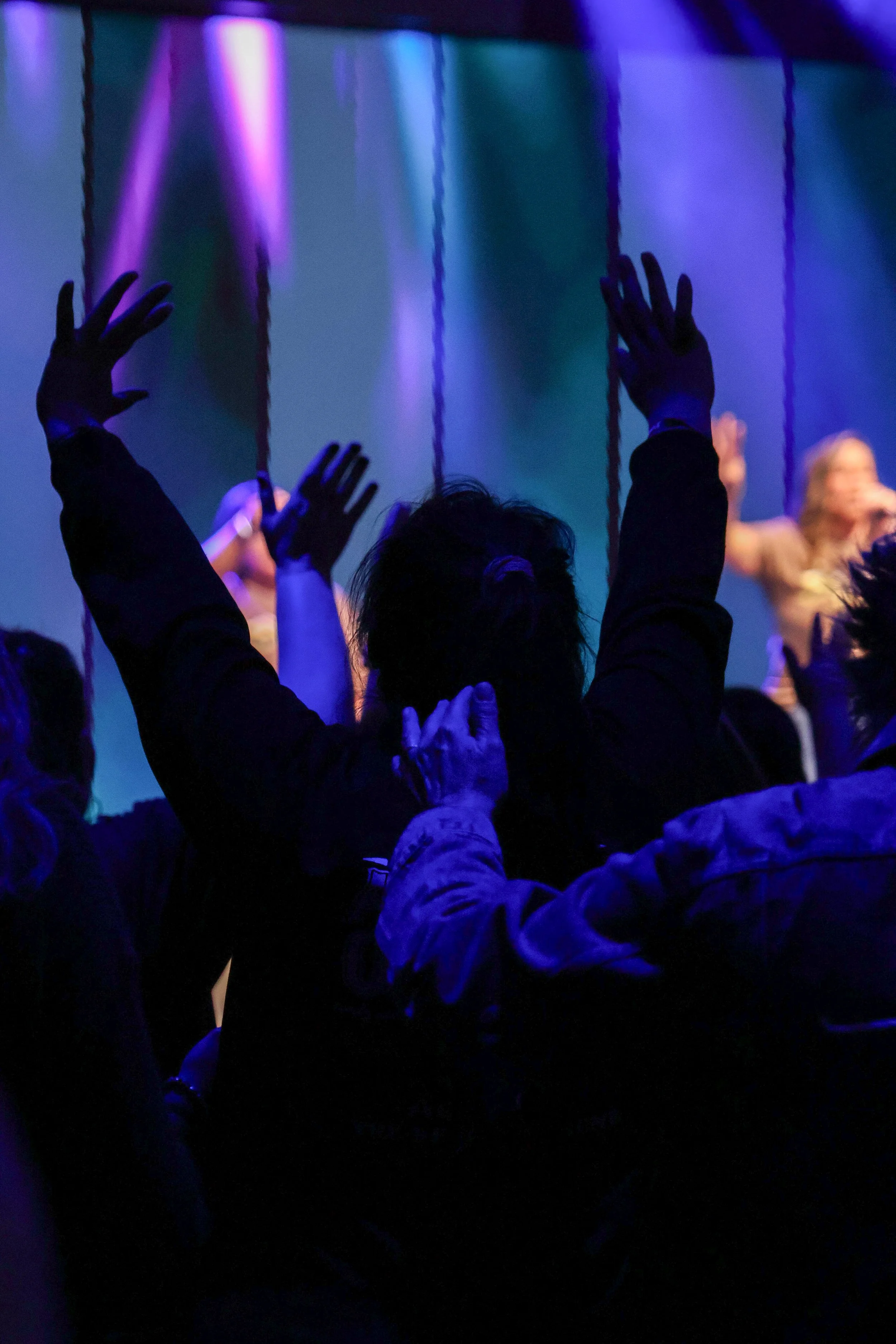 A person in a dark setting with blue and purple lighting, raising both arms above their head with hands open, surrounded by other people, in what appears to be a concert or performance.