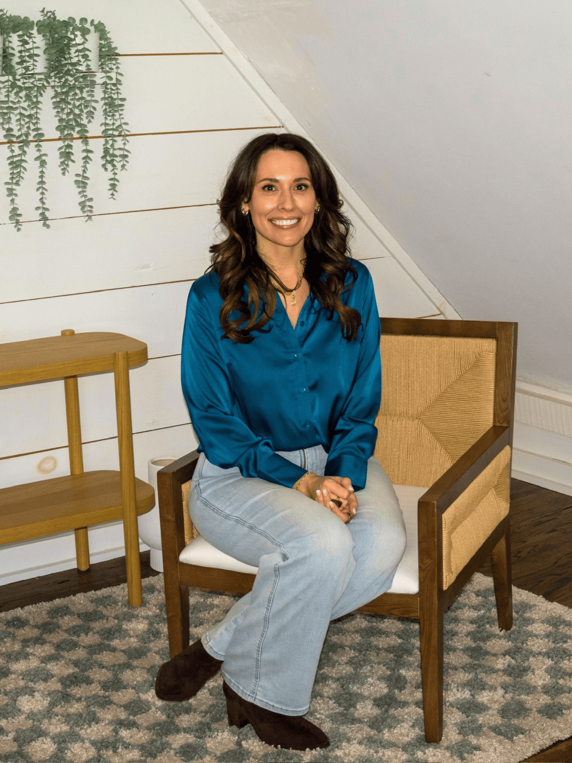 A woman with long, wavy brown hair sitting on a wooden chair with a woven backrest. She is wearing a blue satin blouse, light-wash jeans, and dark boots. The background includes white shiplap walls, a green hanging plant, and a rug with a geometric pattern.