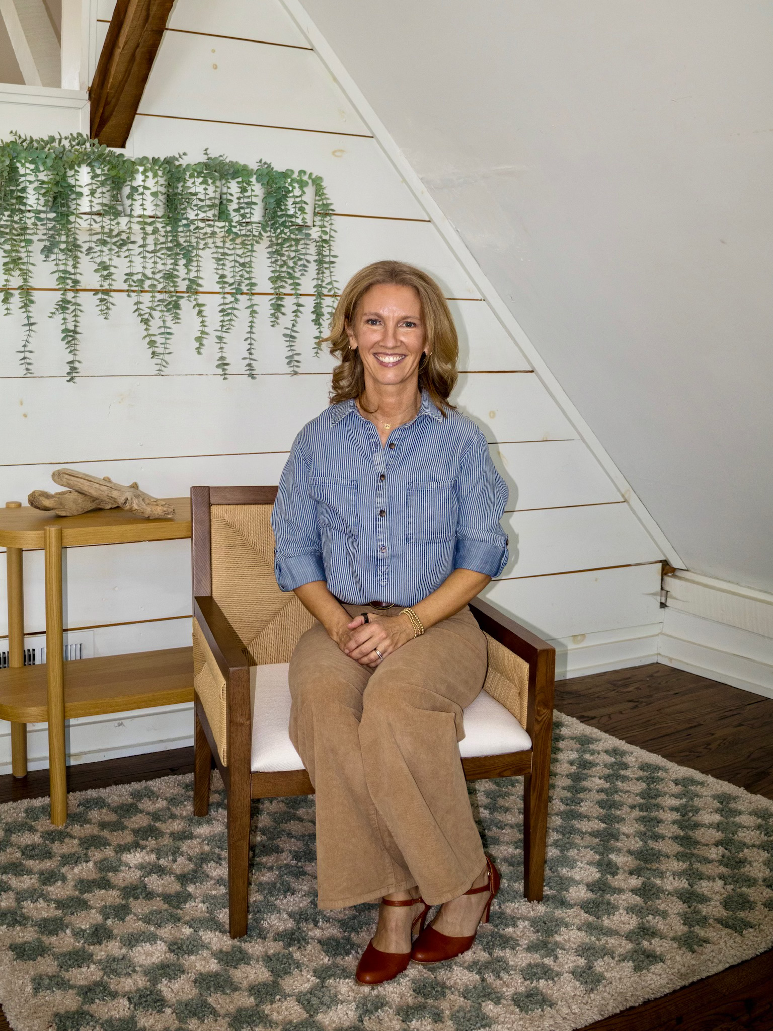 A woman with shoulder-length light brown hair, wearing a blue striped button-up shirt, tan pants, and brown heels, sitting on a wooden chair with a white cushion in a room with white shiplap walls and a patterned rug, smiling at the camera.