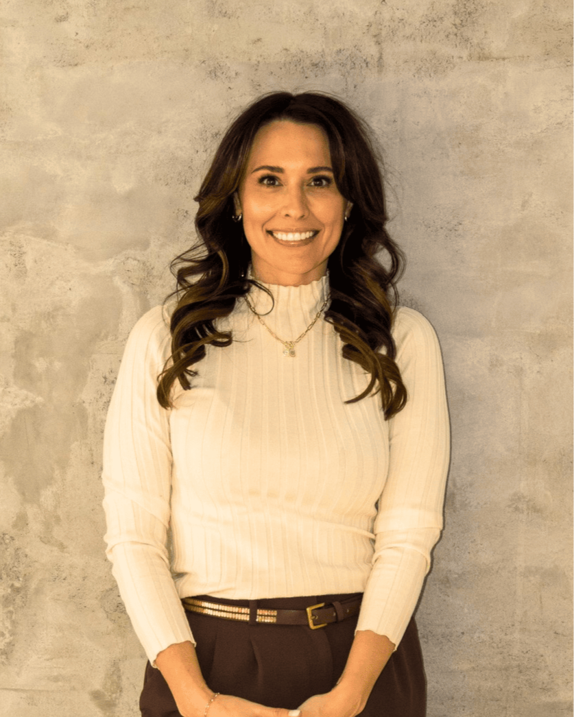 A woman with long, wavy brown hair smiling, wearing a cream-colored ribbed turtleneck sweater and brown pants with a belt, standing in front of a textured beige wall.