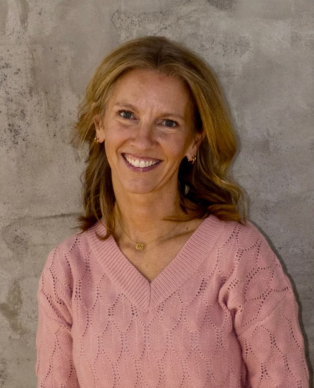 A woman with shoulder-length reddish hair smiling, wearing a pink sweater with a diamond pattern, gold hoop earrings, and a gold necklace with a small pendant, standing against a gray concrete wall.