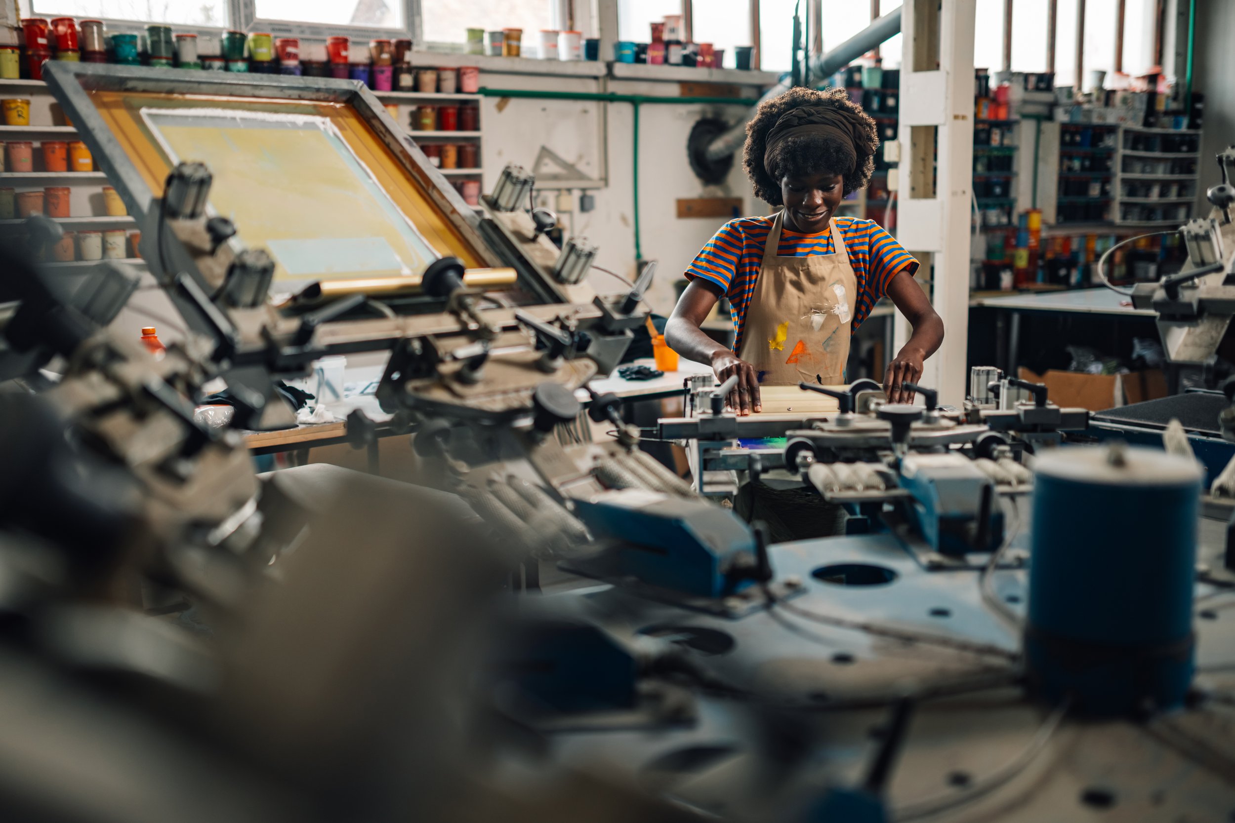 Person in a screen printing workshop, wearing an apron and working with printing equipment, surrounded by colorful paint containers.