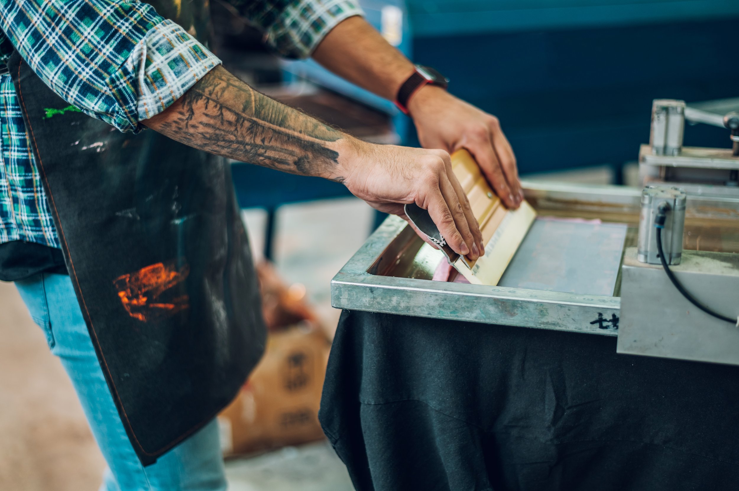 Person screen printing on fabric with a squeegee, wearing an apron and plaid shirt.