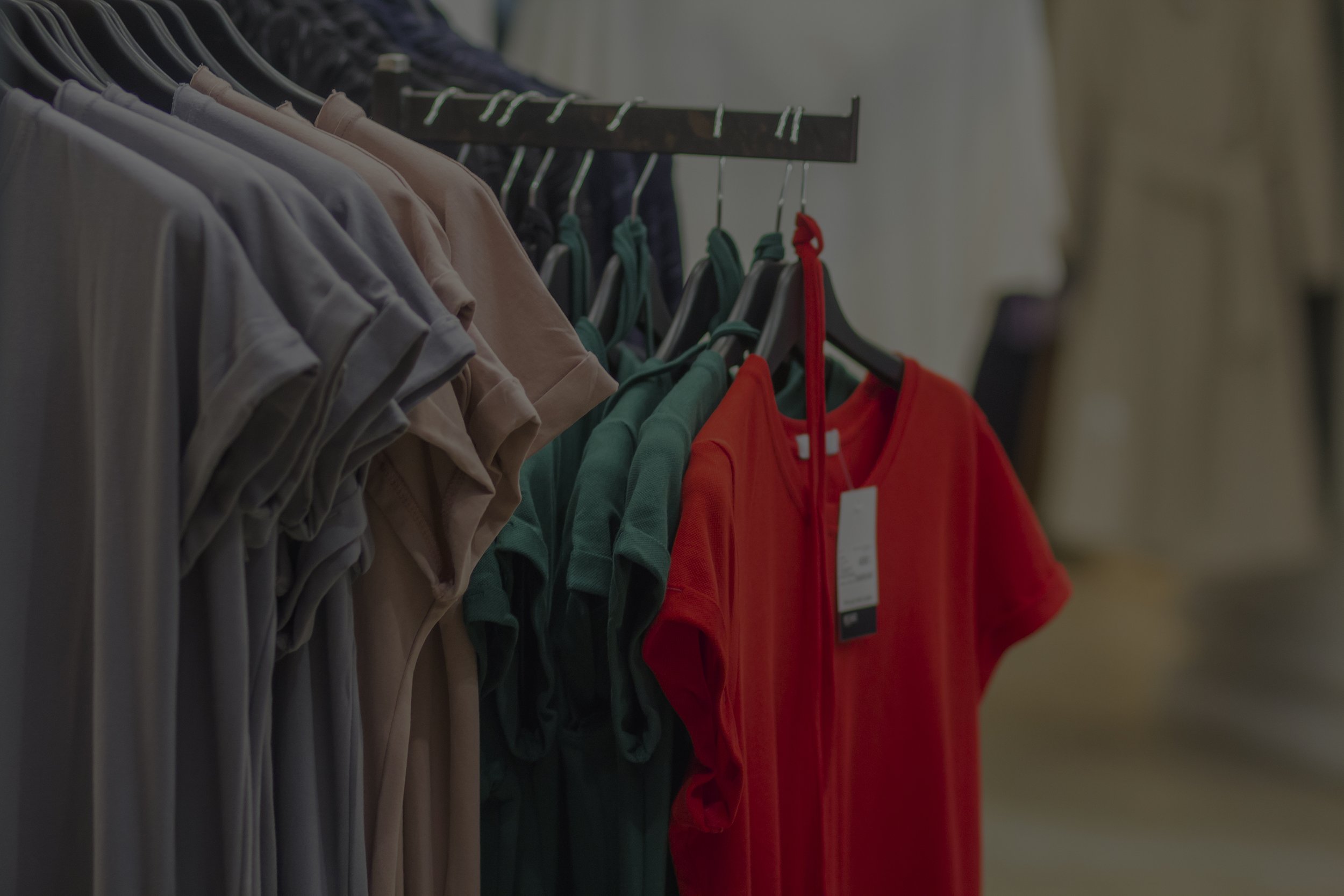 A rack of assorted colored T-shirts on hangers, including gray, beige, green, and a prominent red shirt with a price tag, in a clothing store.