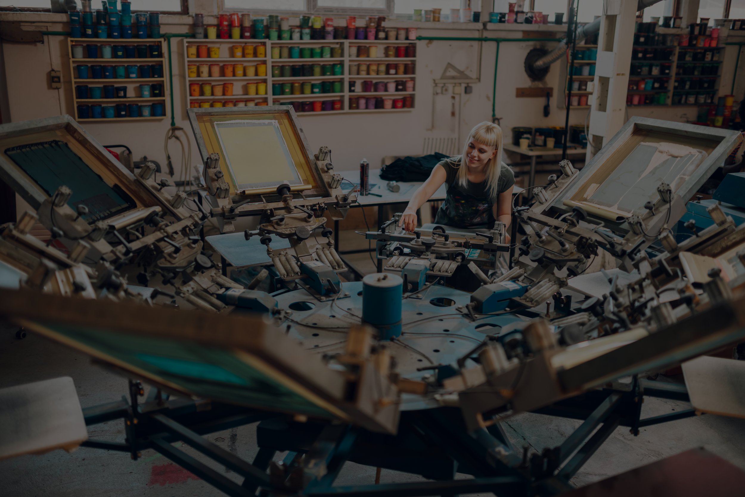 Person operating a screen printing machine in a studio with colorful shelves of ink.