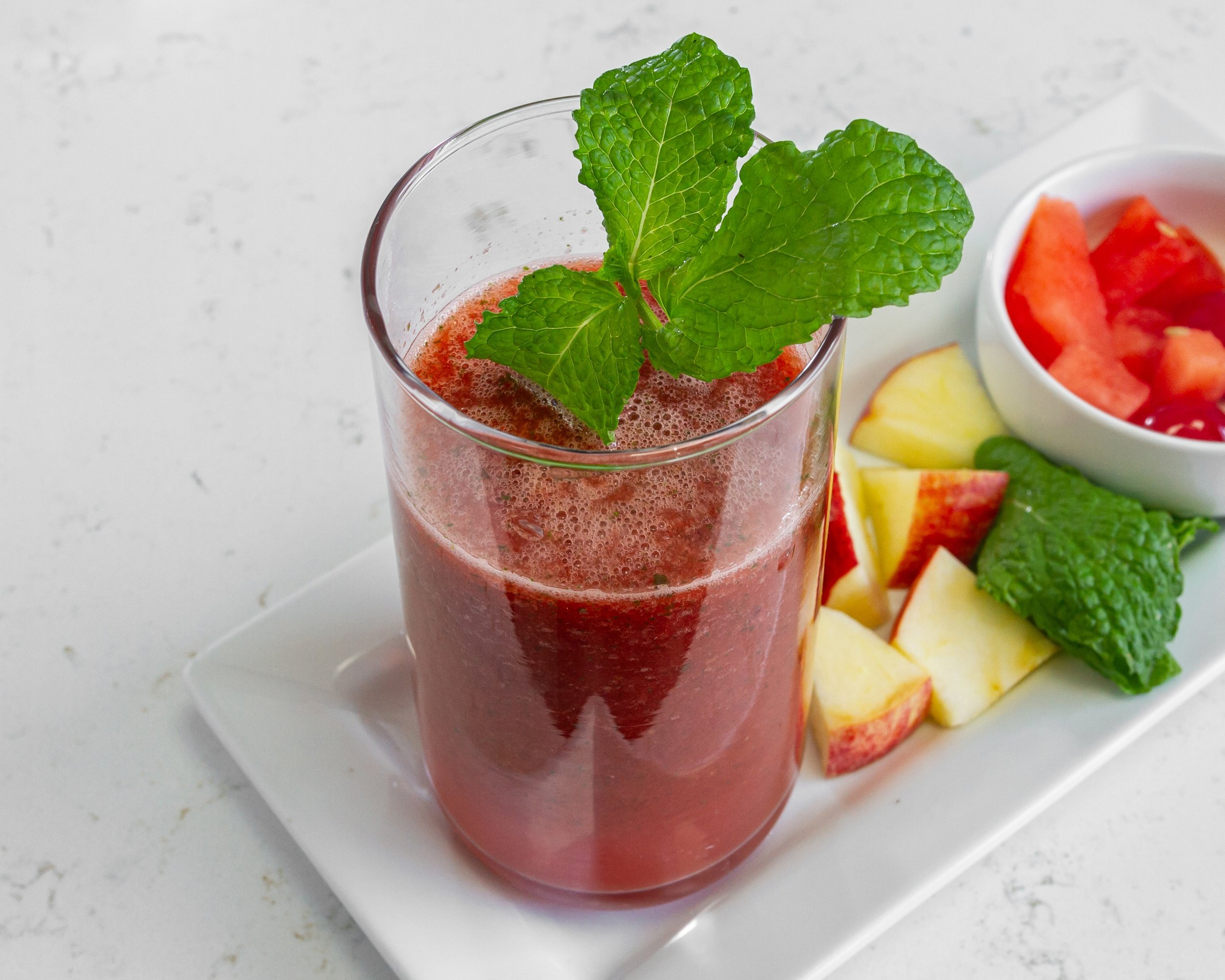 Glass of red juice with mint garnish on white tray, surrounded by chopped apple pieces, mint leaves, and a small bowl of tomato pieces.