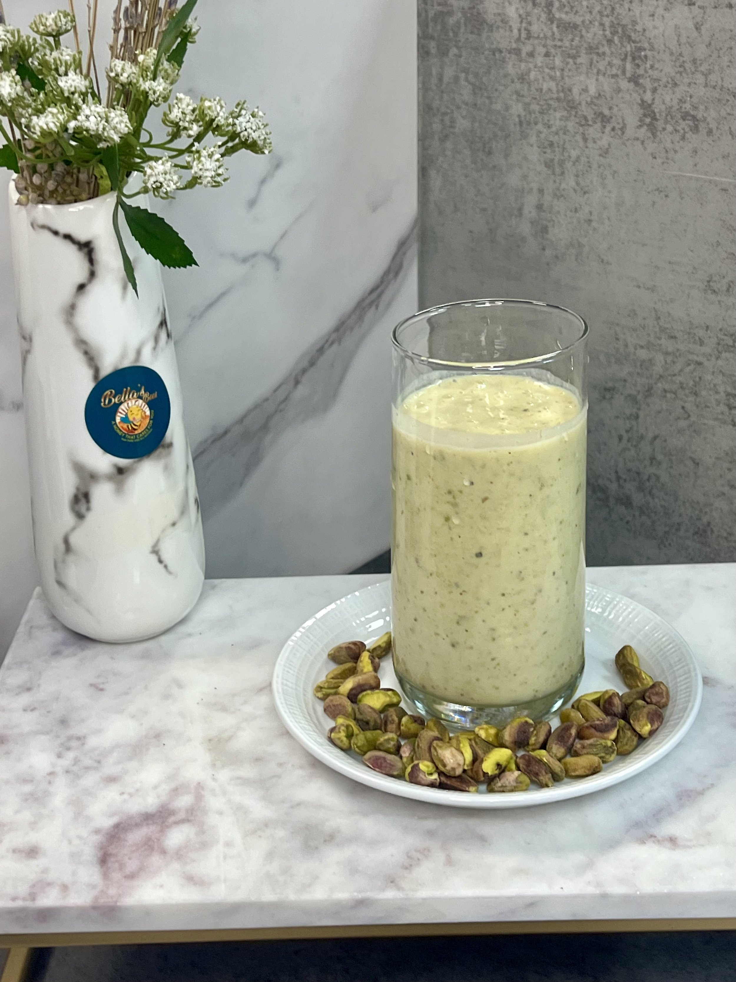 A glass of pistachio smoothie on a white plate with pistachios, next to a marble vase with flowers, on a marble countertop.