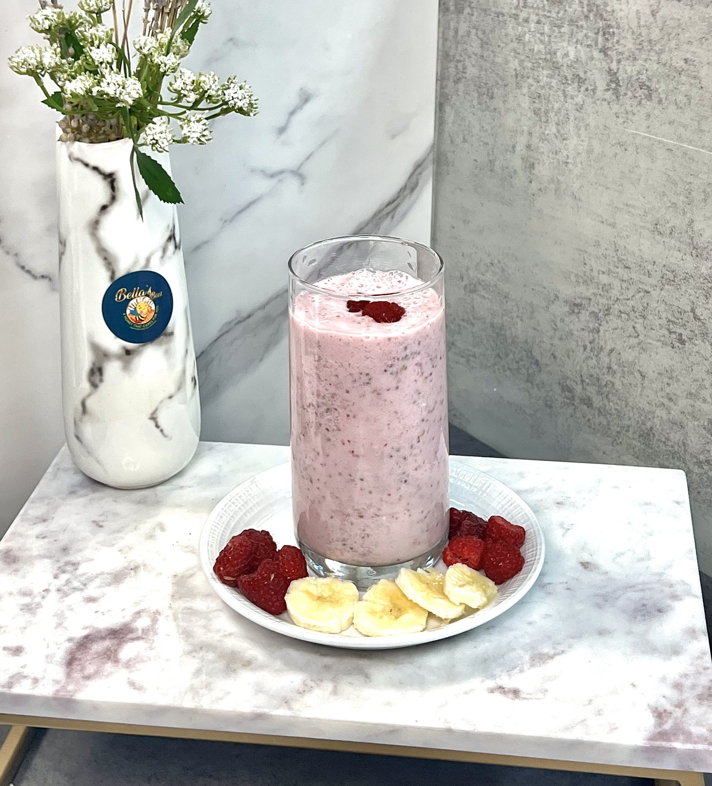 A glass of pink smoothie on a plate surrounded by raspberries and banana slices, set on a marble table next to a white marble vase with flowers.