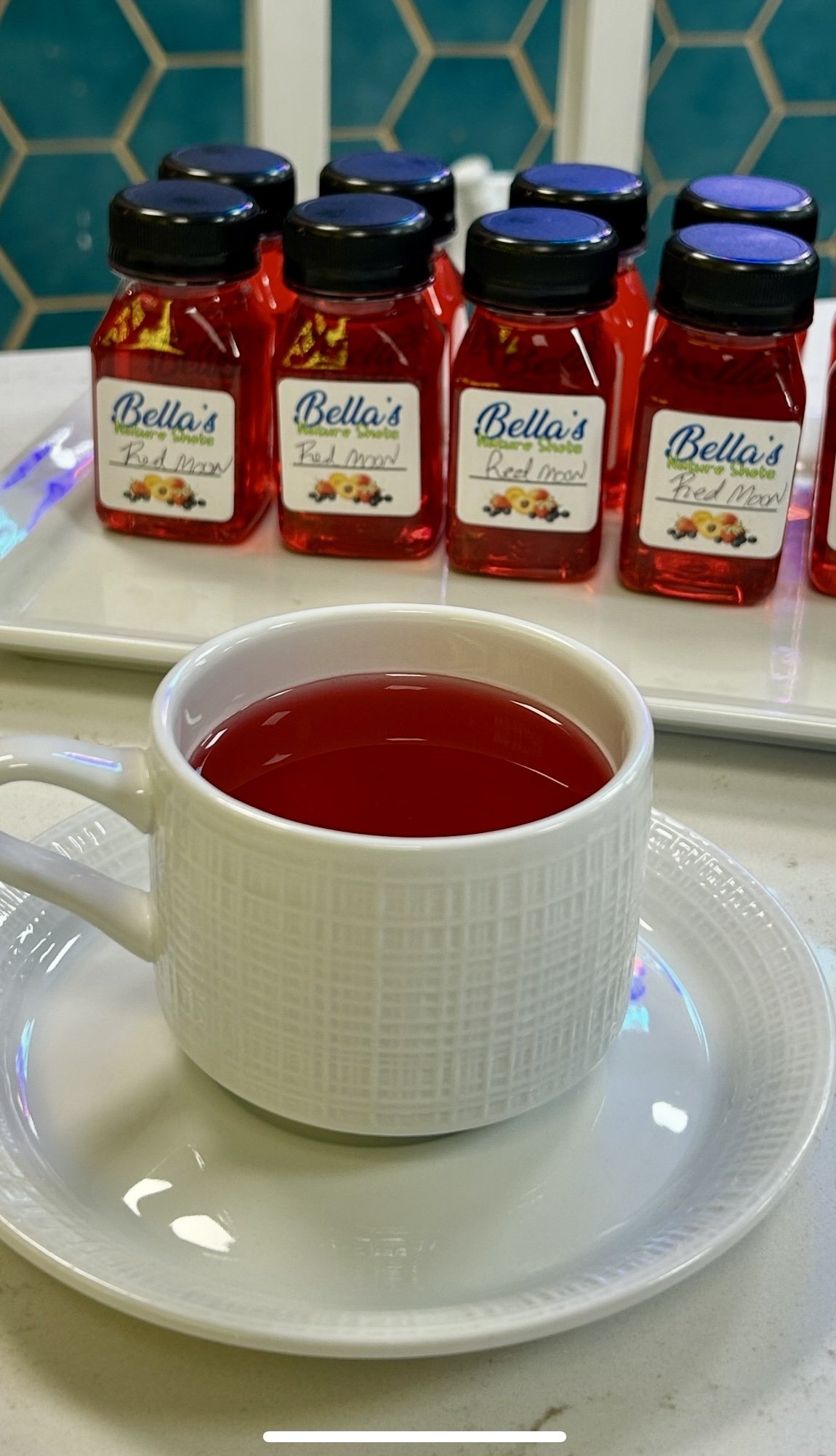 A white cup with red liquid on a saucer, in front of bottles labeled 'Bella's Red Moon.'