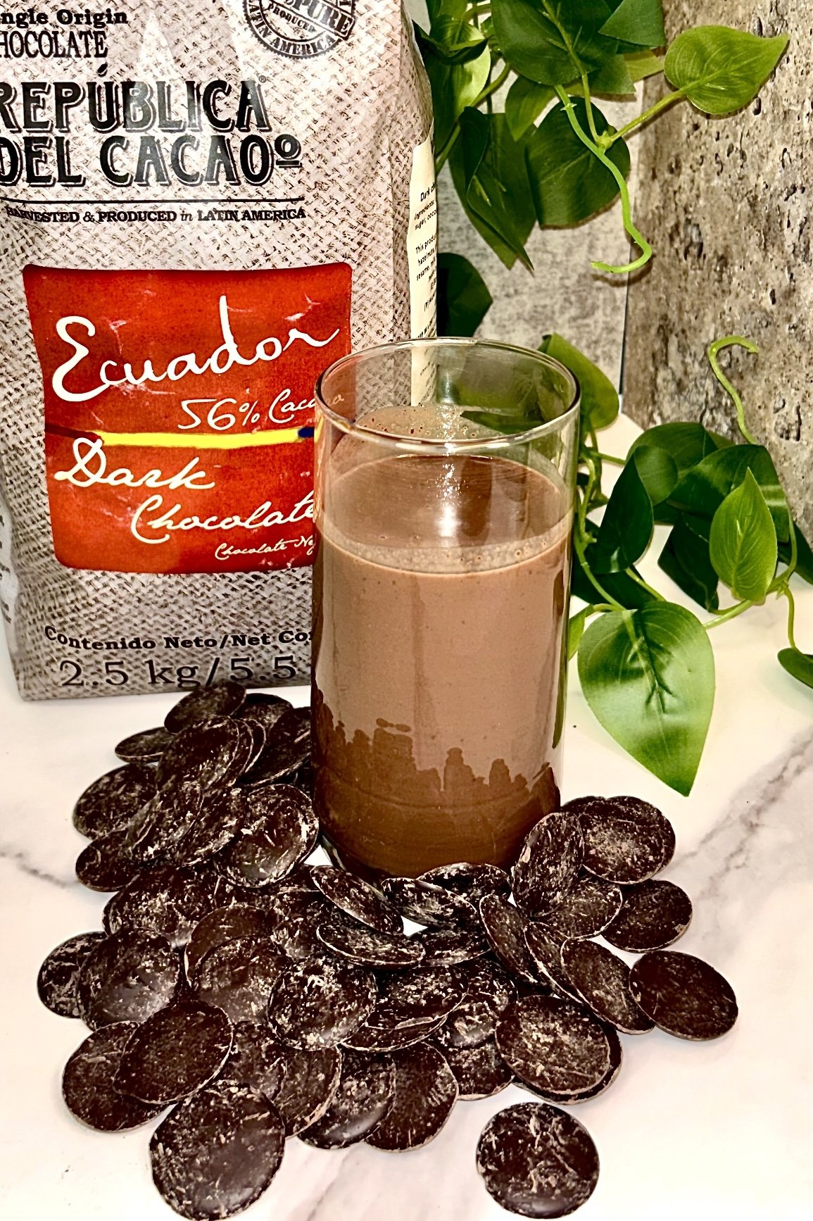 Dark chocolate products with Ecuador label, a glass of chocolate drink, chocolate discs scattered, and green plant leaves in the background.