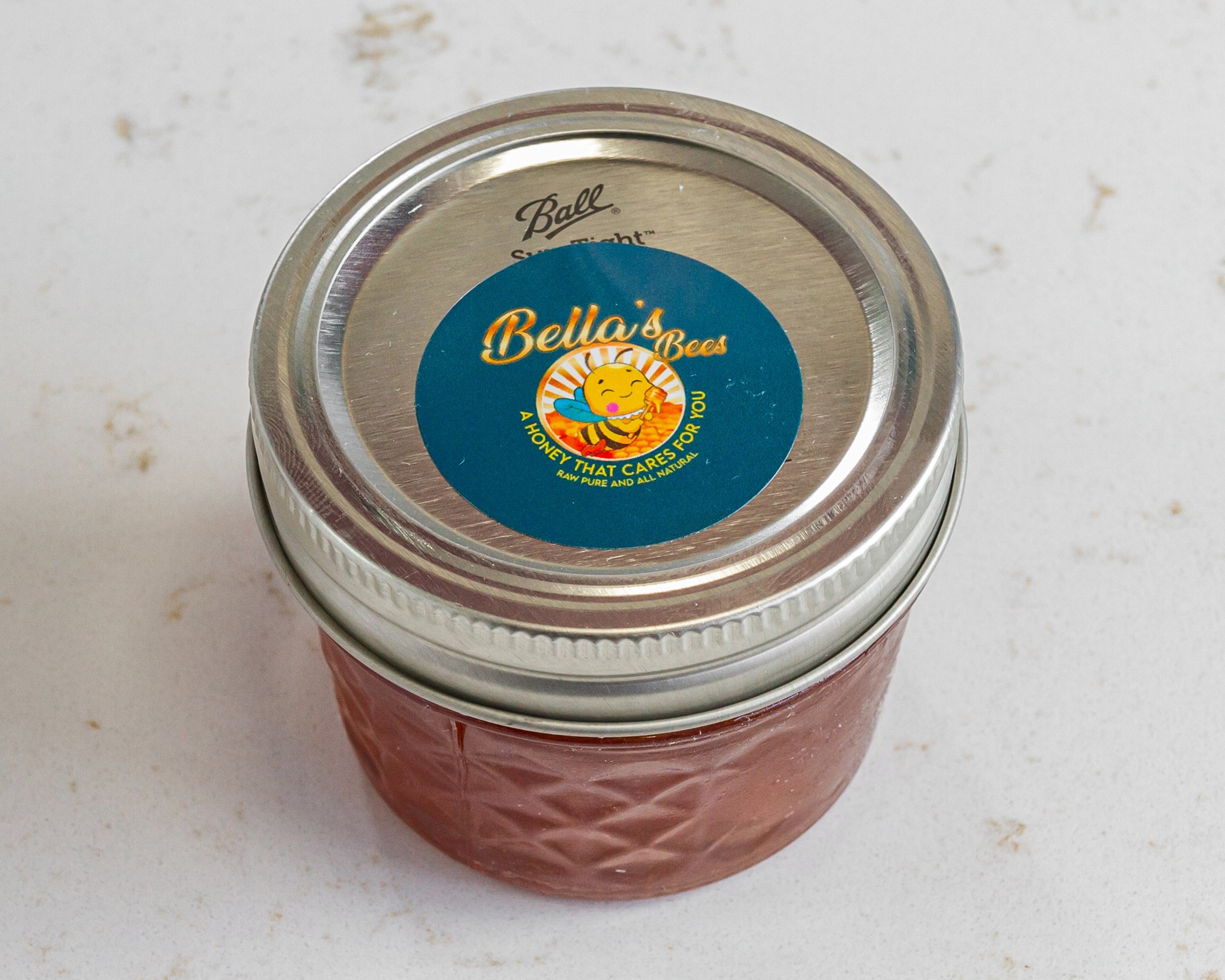 Jar of honey with a label on the lid reading 'Bella's Bees, A Honey That Cares for You'