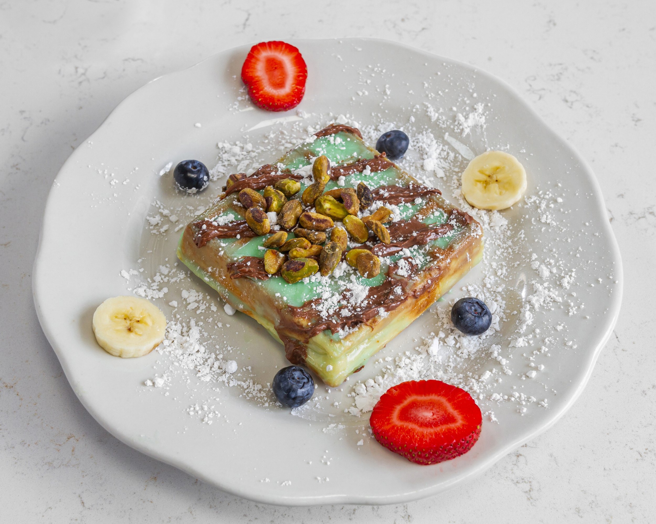 Dessert plate with pistachio-topped cake, garnished with sliced strawberries, bananas, blueberries, and powdered sugar.