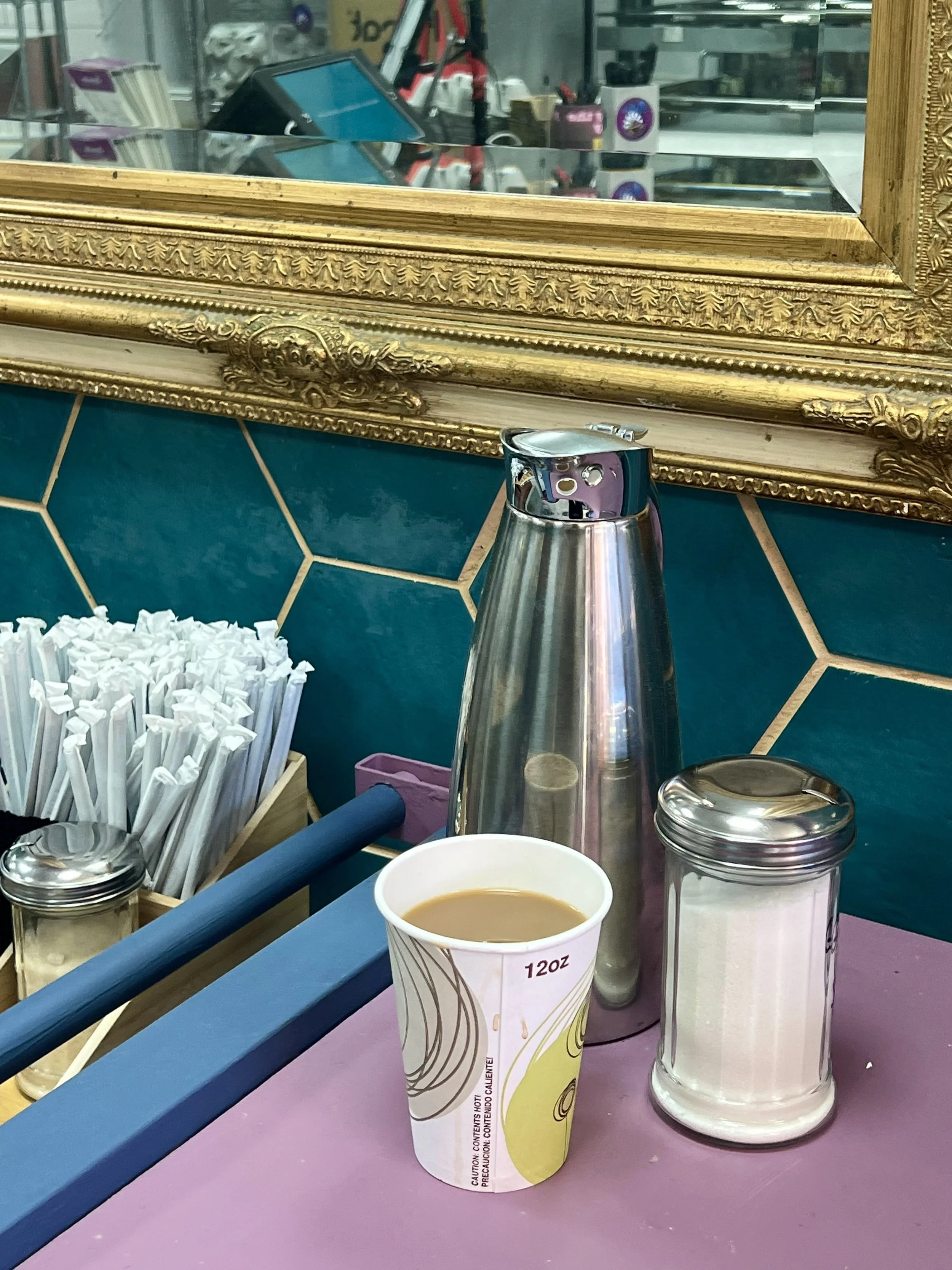 A paper coffee cup with a beverage, a sugar dispenser, and a metal thermos are on a countertop, in front of a decorative framed mirror and tile wall.