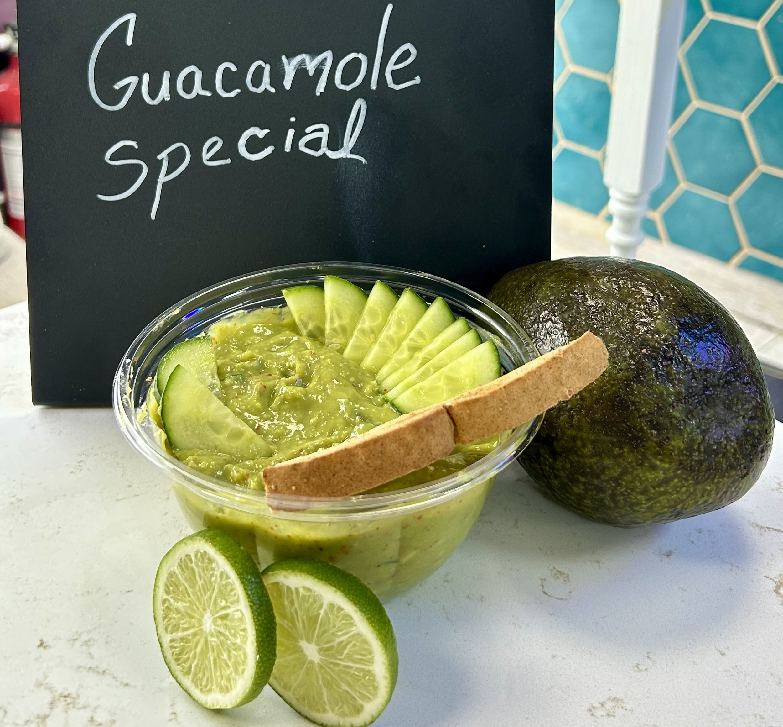 A bowl of guacamole garnished with cucumber slices and a breadstick, placed next to an avocado and sliced lime on a table. A sign behind it reads 'Guacamole Special'.