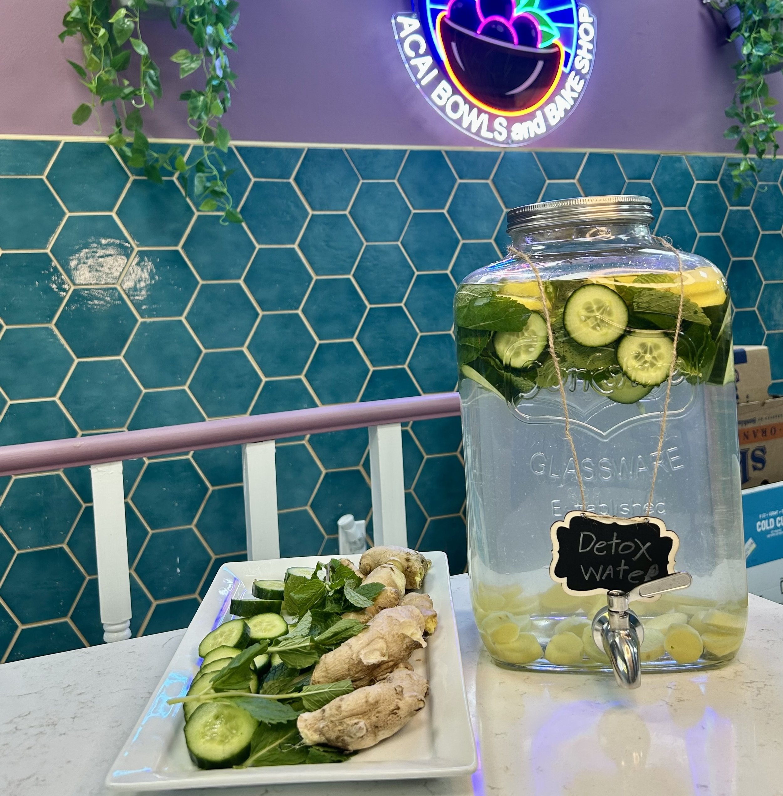 Glass container of detox water with cucumbers, lemons, and mint on a counter, next to a plate of ginger, cucumber slices, and mint leaves. Background with teal hexagonal tiles and neon sign.