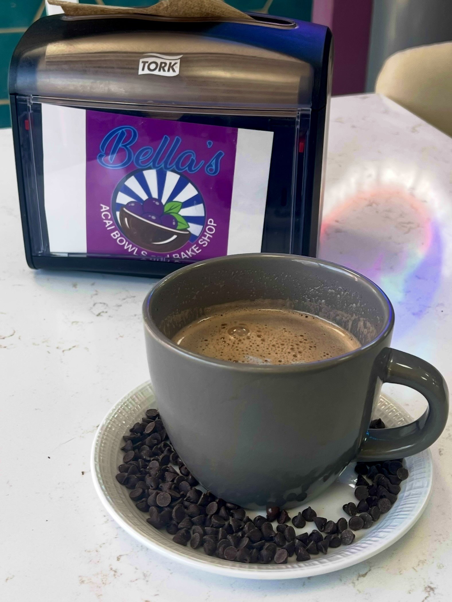 A gray mug filled with frothy hot chocolate on a white saucer surrounded by chocolate chips. In the background, a metal napkin dispenser with a sign reading "Bella's Acai Bowls and Bake Shop."