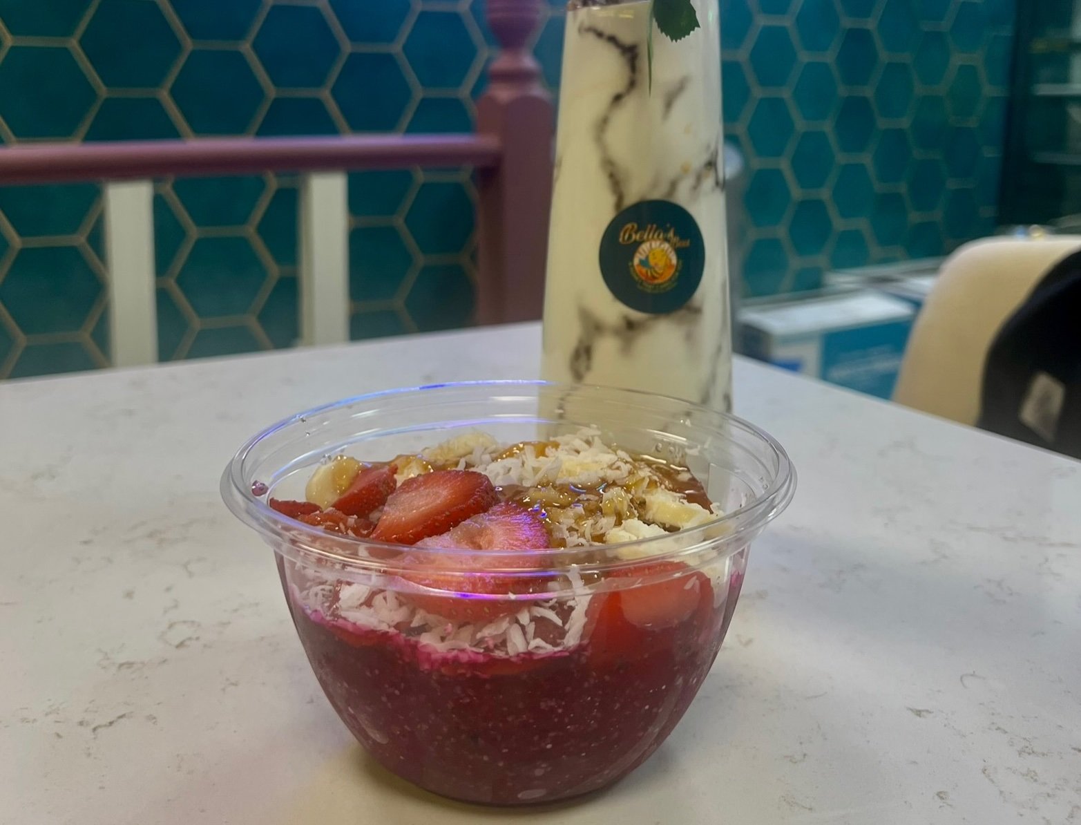 Acai bowl topped with strawberries, coconut flakes, and granola, placed on a table beside a marble-designed bottle.