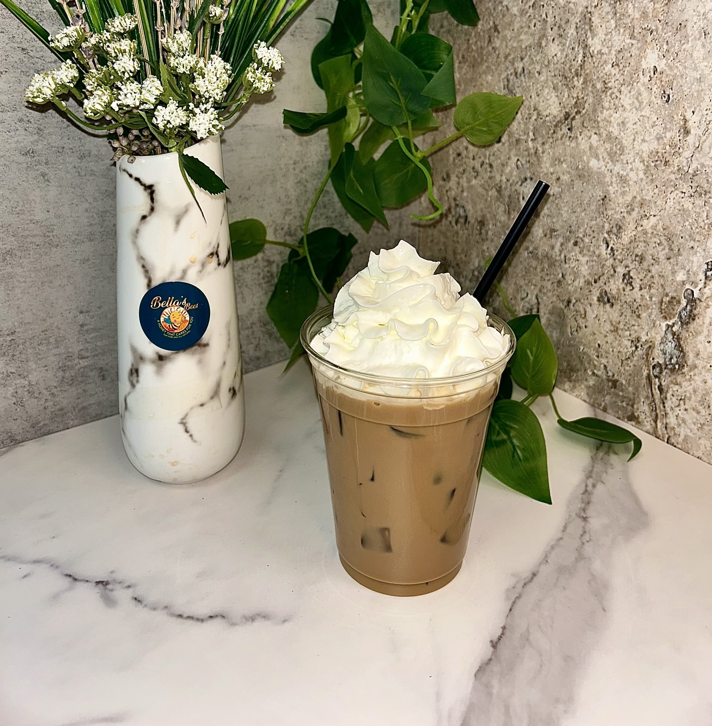 Iced coffee with whipped cream and straw next to a marble-effect vase with flowers and greenery on a marble surface.