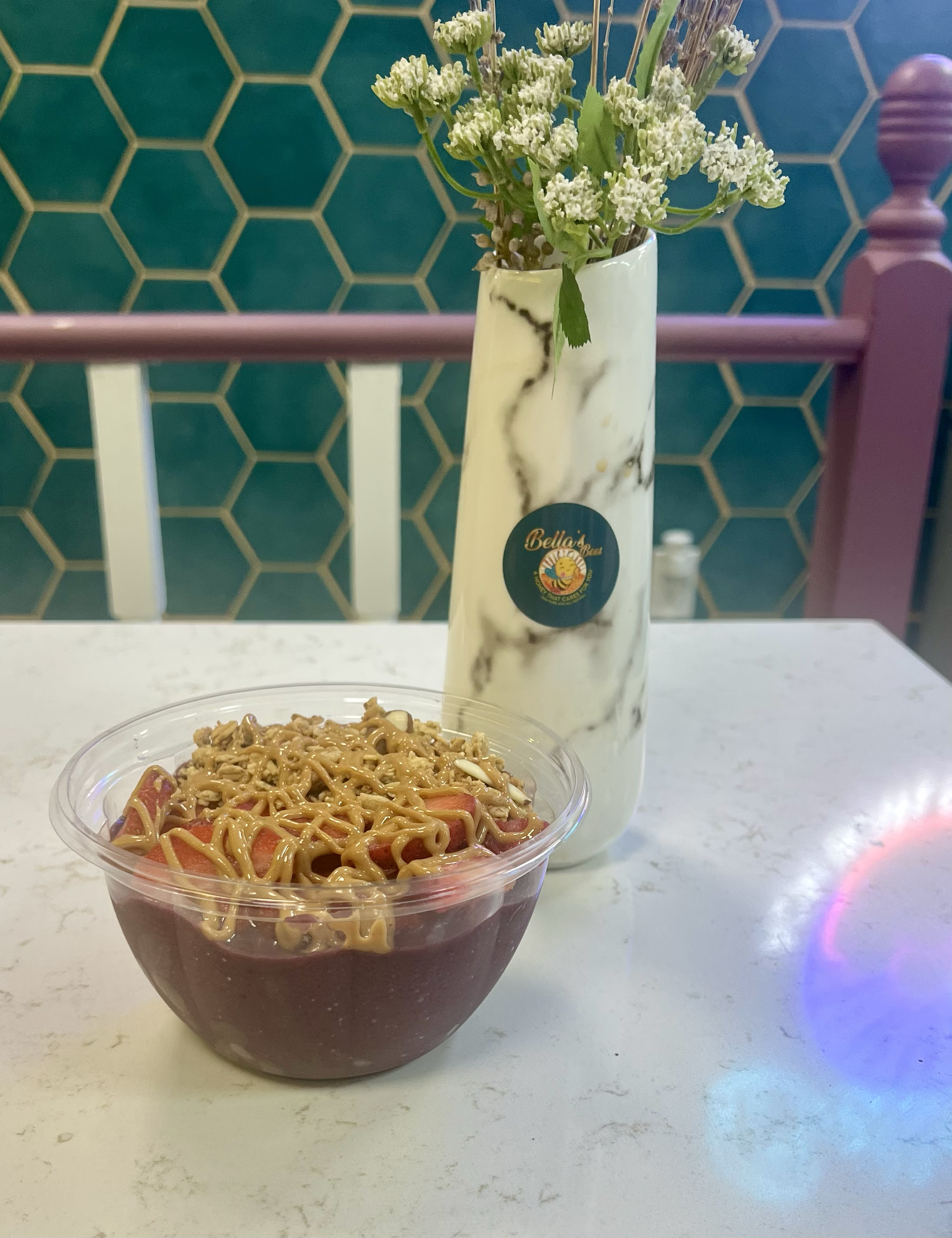 Acai bowl topped with sliced fruit and nut butter, next to a decorative vase with flowers, placed on a white table with a hexagonal green tiled background.