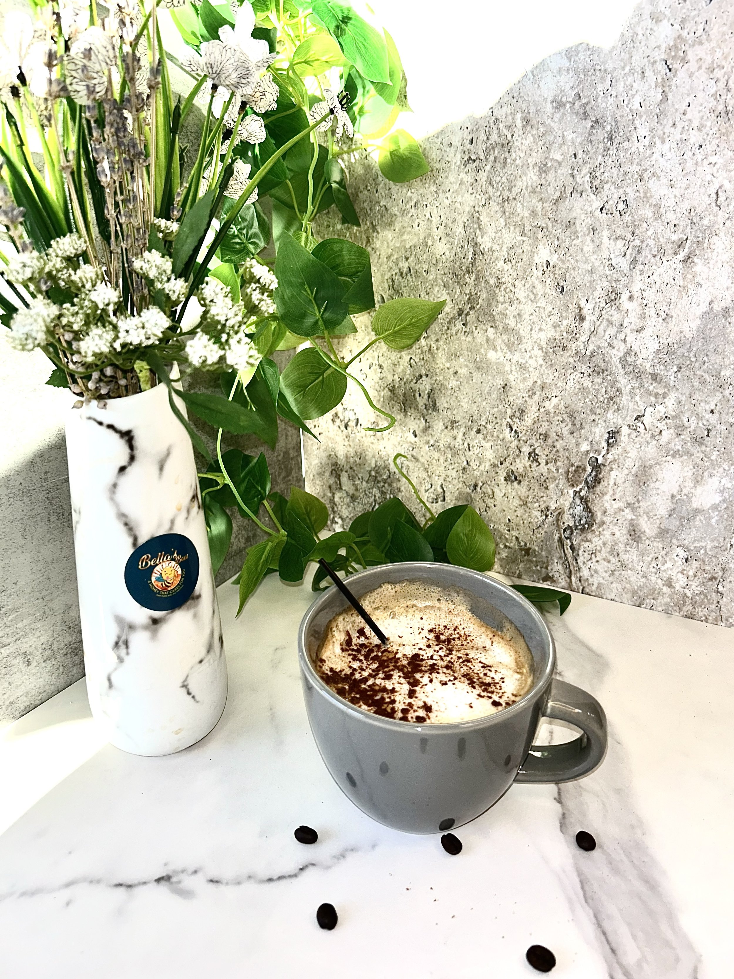 A gray mug of cappuccino with cocoa powder on top sits on a marble surface. Coffee beans are scattered nearby. A marble-textured vase with a label holds a bouquet of white flowers and green foliage. A textured stone backdrop is in the background.