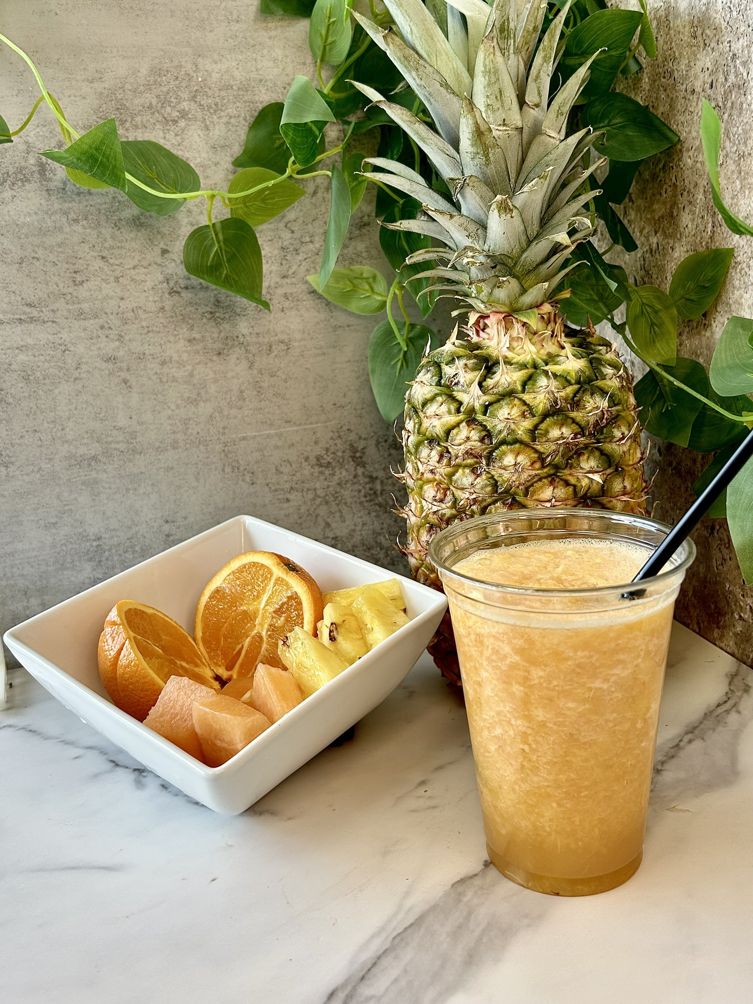Fresh fruit smoothie in a plastic cup with a straw, next to a whole pineapple and a bowl of sliced orange, pineapple, and grapefruit on a marble surface, with green leaves in the background.
