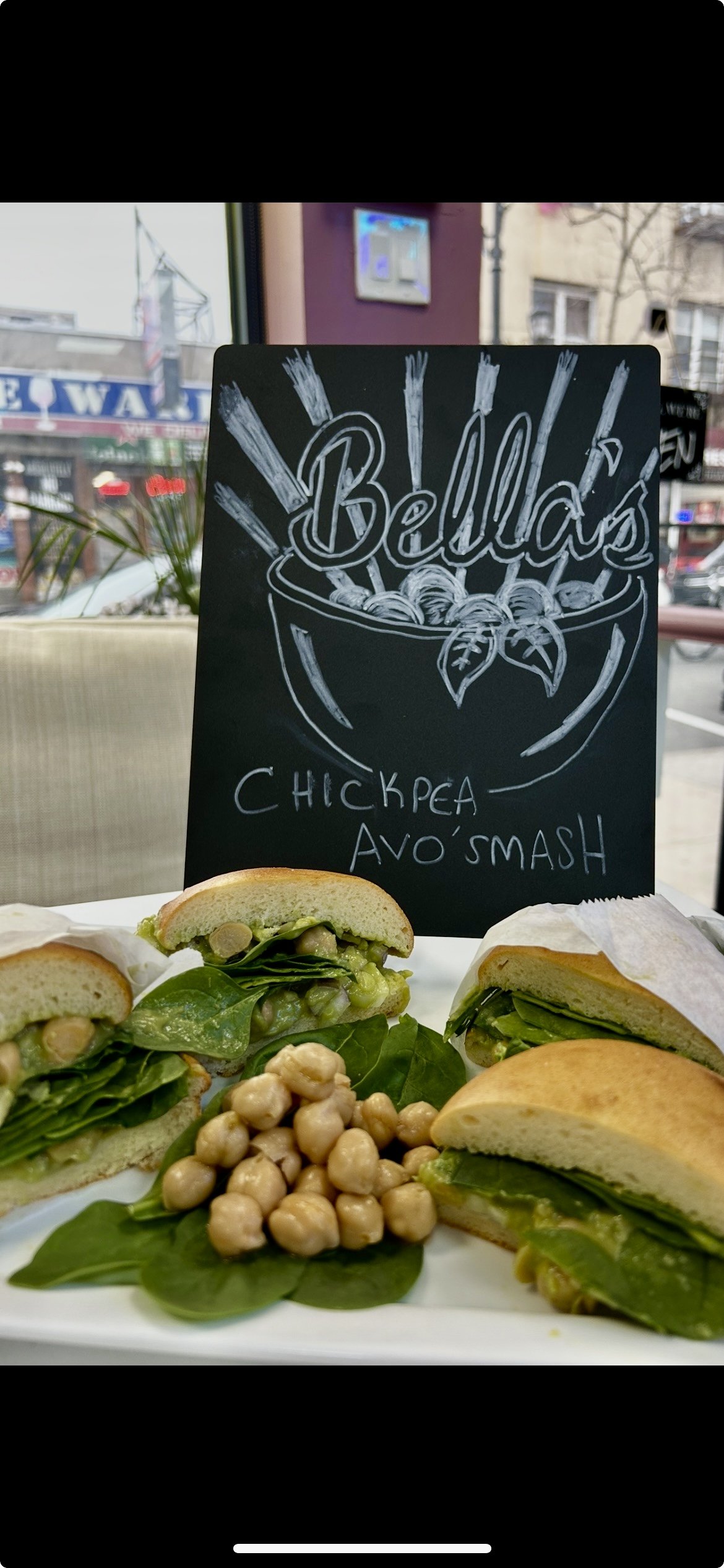 Chalkboard sign with 'Bella's' and 'Chickpea Avo Smash' written on it, with sandwiches containing chickpeas, avocado, and greens on a plate.