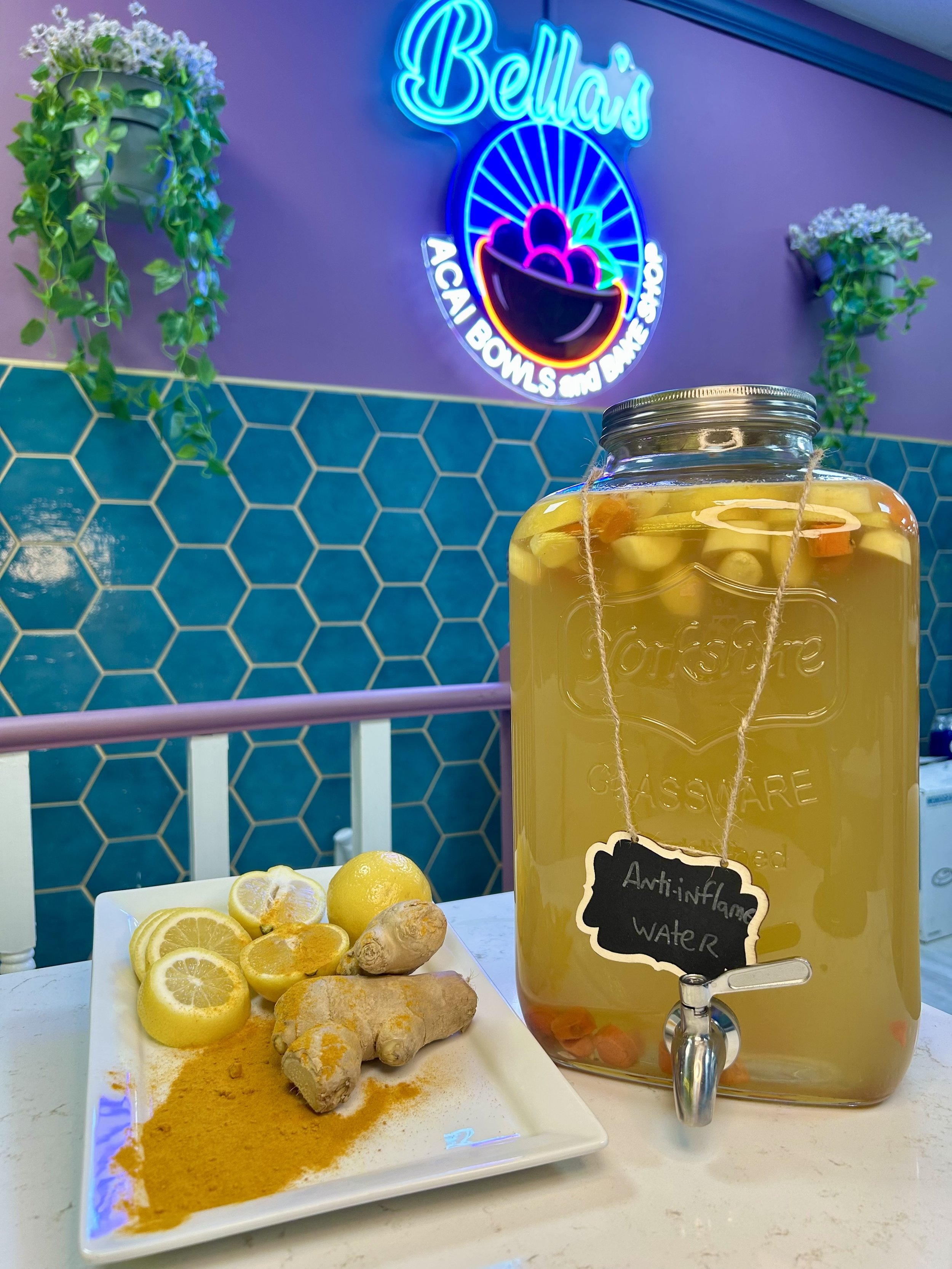 A display of anti-inflammatory water in a glass dispenser with lemons, ginger, and turmeric on a plate, against a background with hexagonal blue tiles and a neon "Bella's Acai Bowls and Bake Shop" sign.