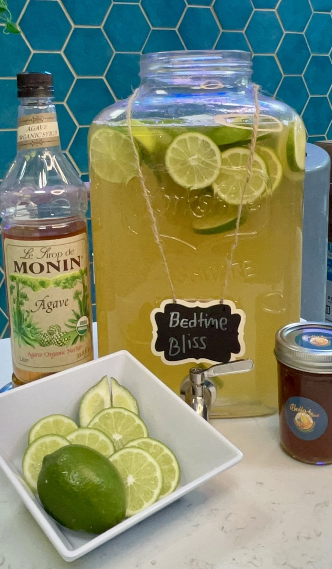 A glass beverage dispenser labeled "Bedtime Bliss" filled with a lime-infused drink, a bottle of organic agave syrup, a bowl of lime slices, and a jar of honey on a counter.