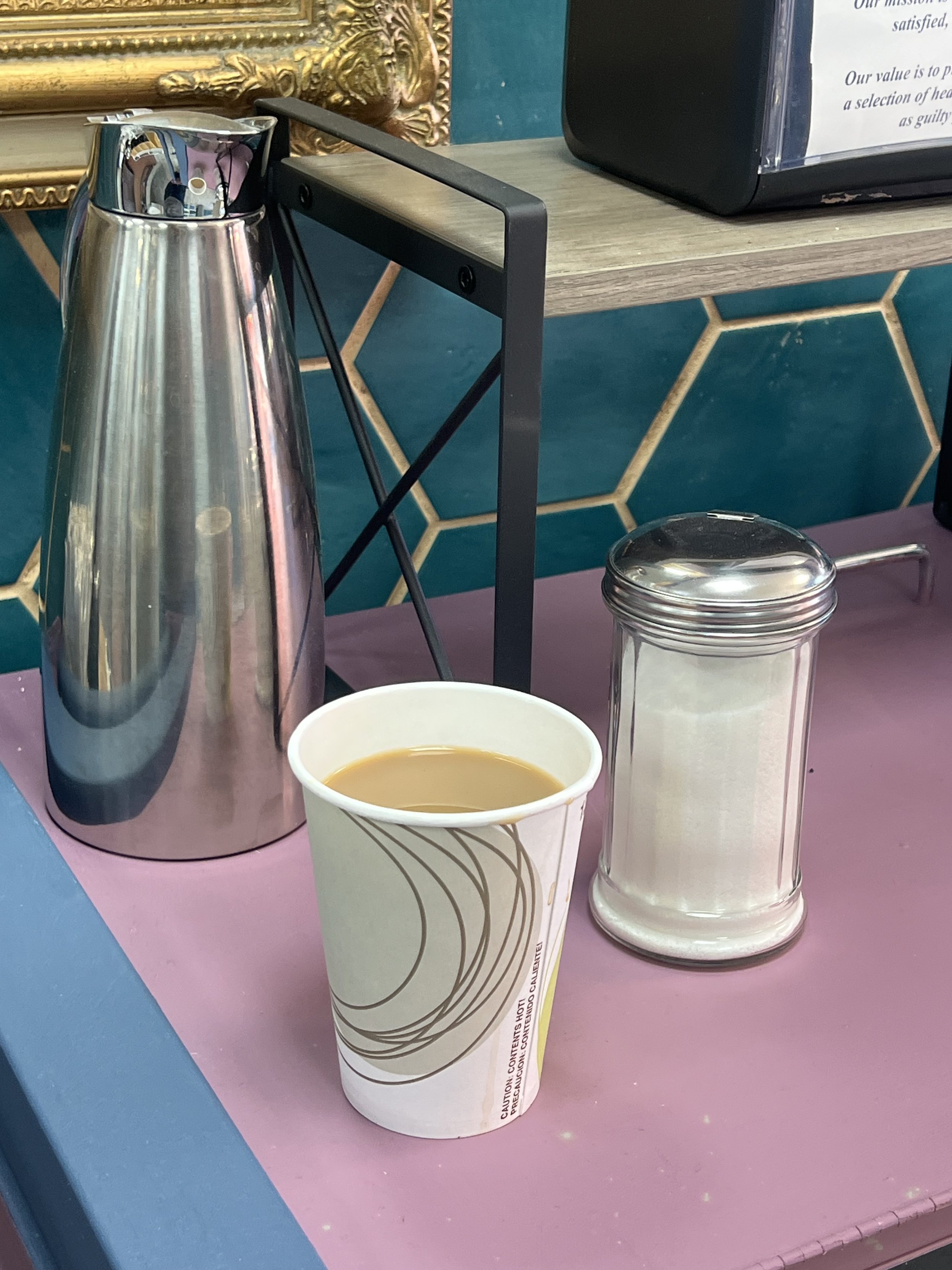 Coffee station with a paper cup filled with coffee, a stainless steel thermos, and a sugar dispenser on a countertop, against a blue hexagonal tile background.