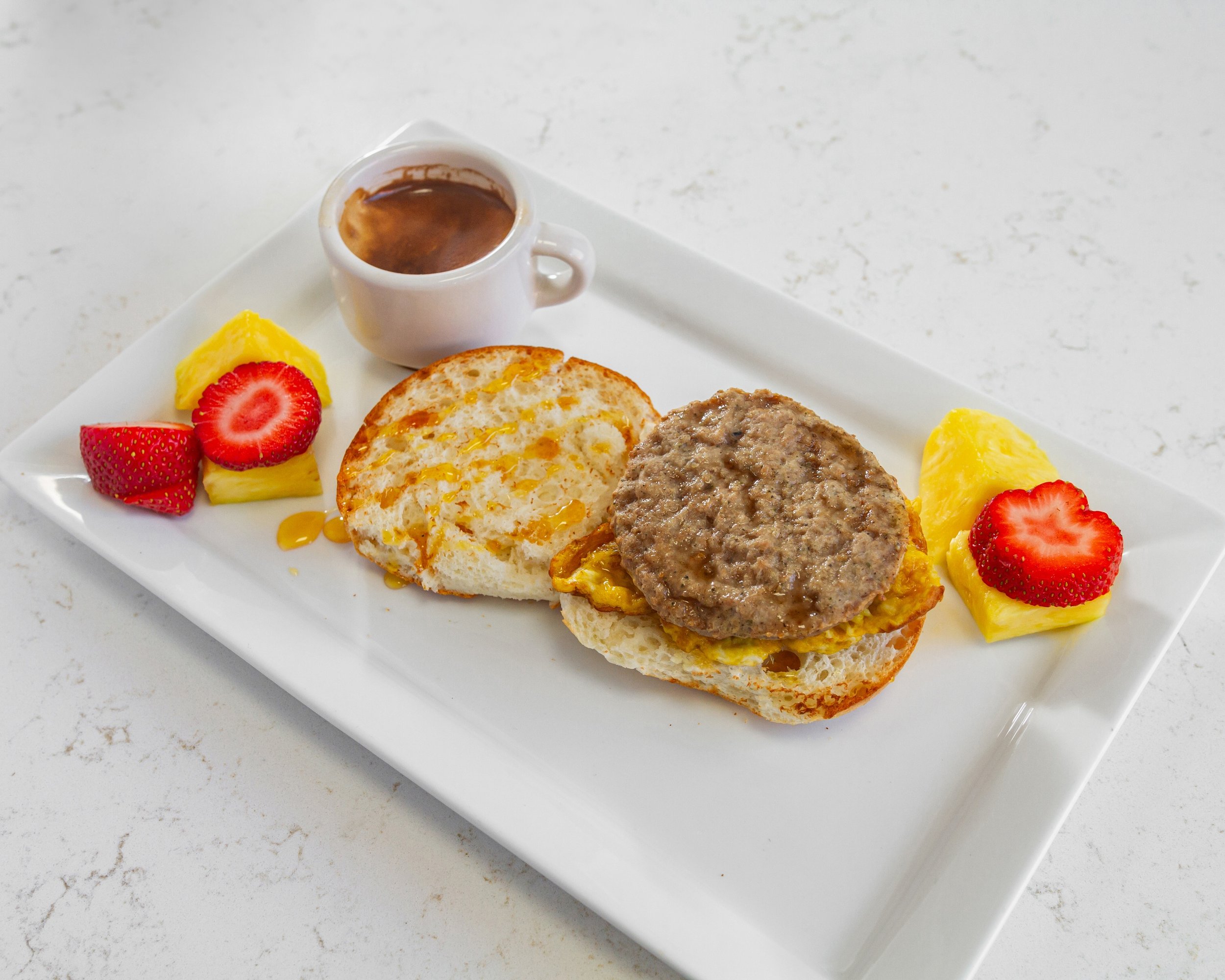 Breakfast sandwich with sausage patty, egg, and English muffin, served with fruit slices and a cup of coffee on a white plate.
