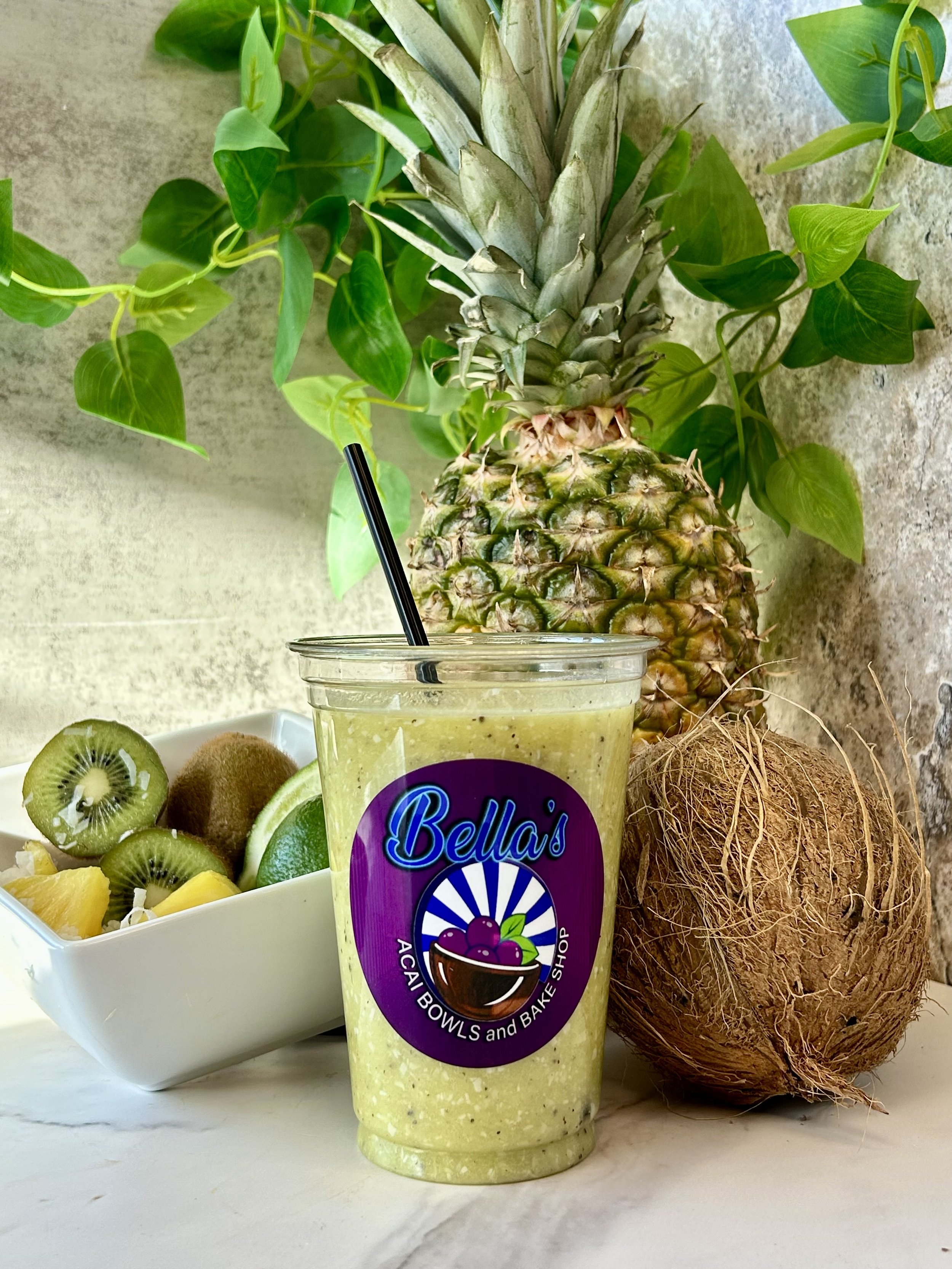 Fresh green smoothie in a clear cup with "Bella's" logo, surrounded by a whole pineapple, coconut, and a bowl of sliced kiwi and other fruits, against a leafy background.