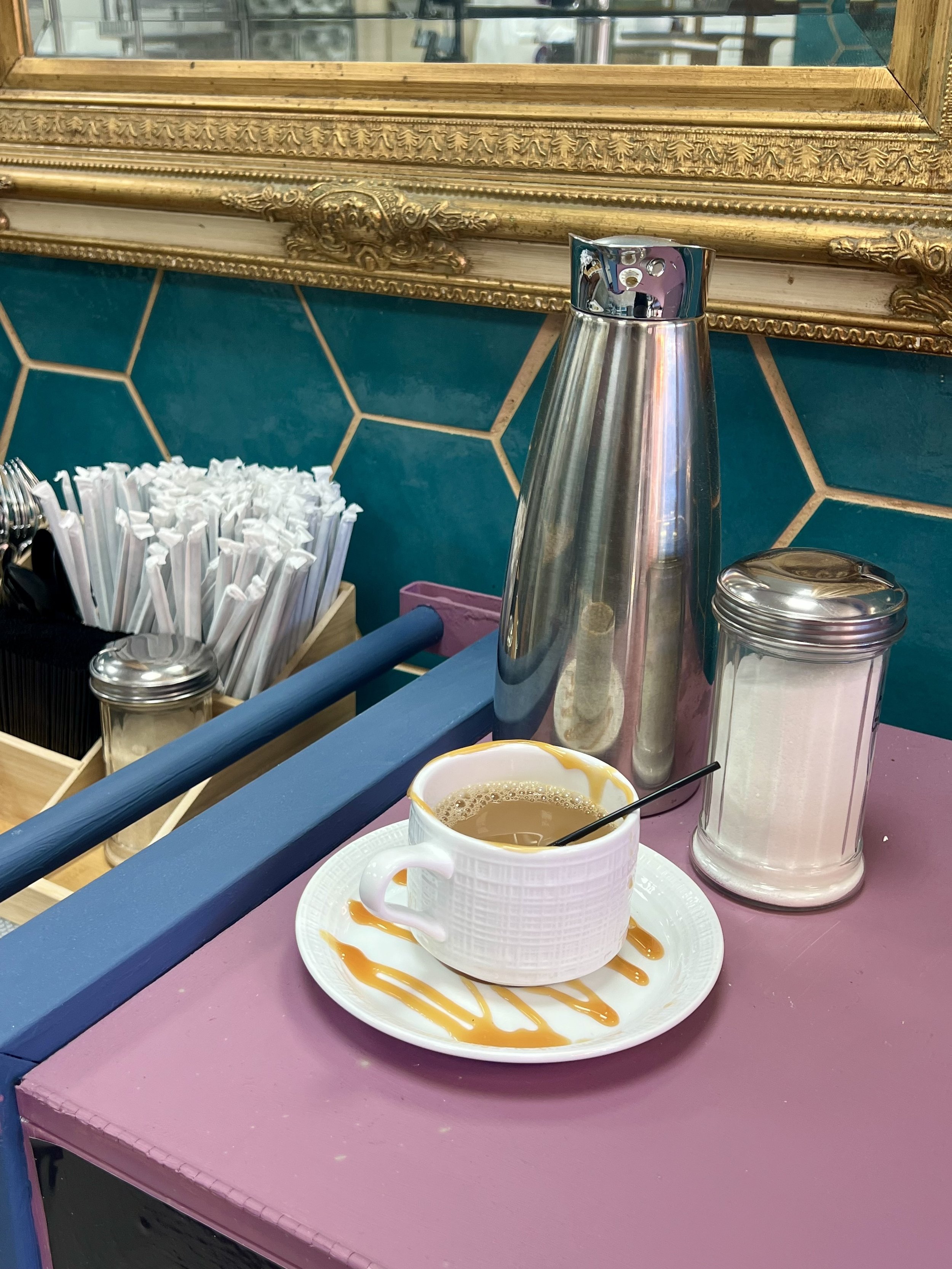 Coffee setup with cup on a saucer, a sugar dispenser, and a metal thermos, set on a purple counter with a decorative mirror and blue hexagonal tiles in the background. Straws and stirrers are also visible.