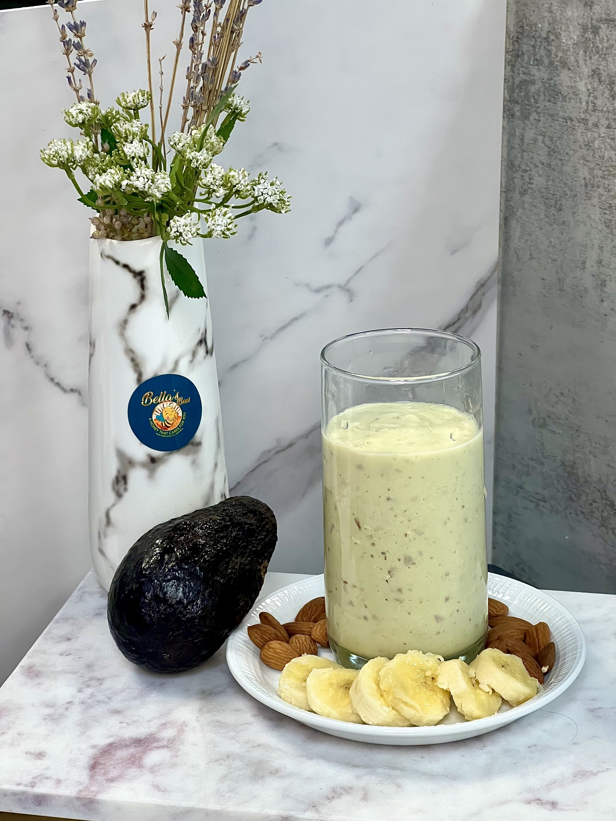 A creamy avocado smoothie in a glass on a plate with sliced bananas and almonds, next to an avocado and a white vase with flowers.