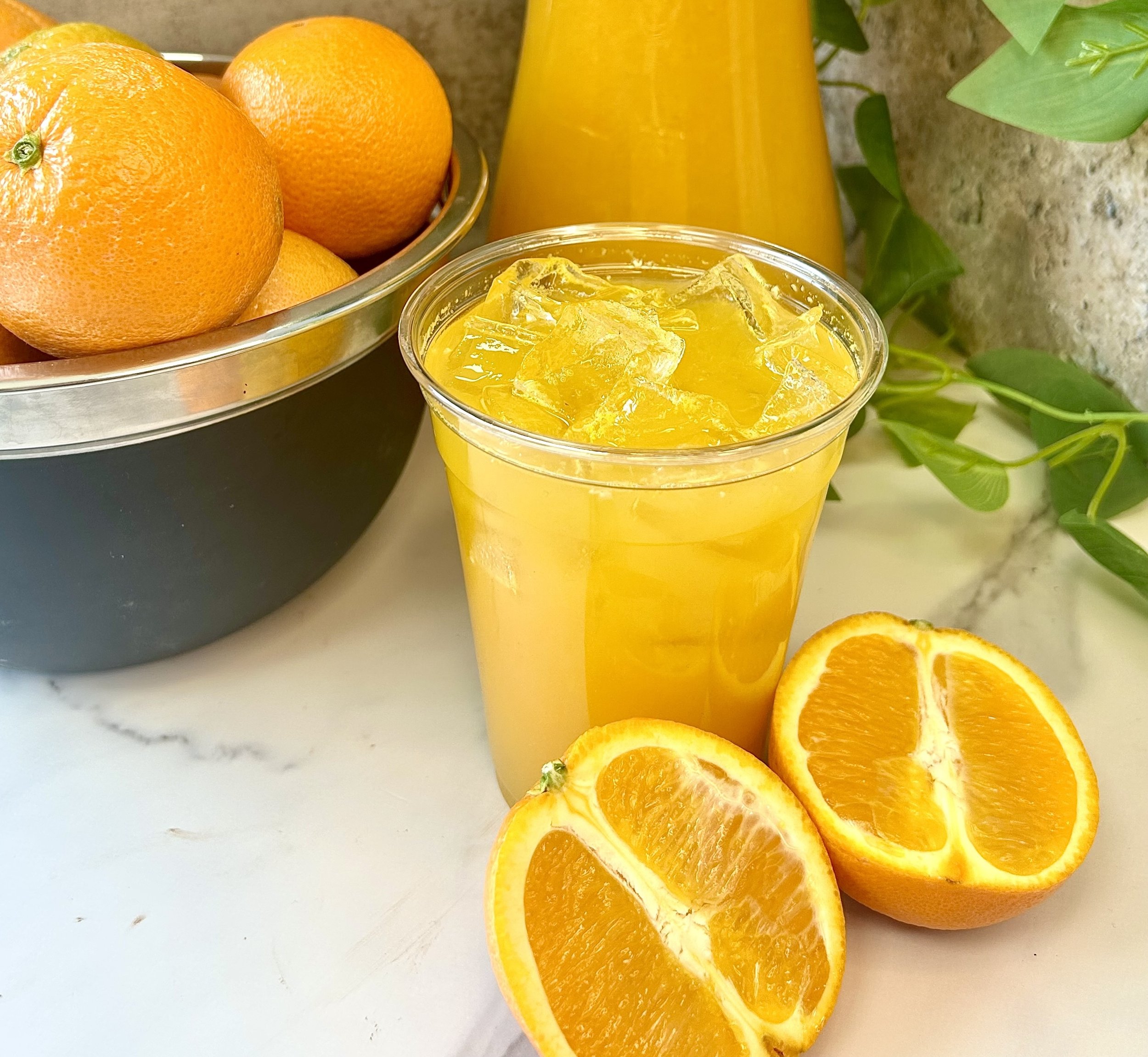 Glass of iced orange juice next to a bowl of whole oranges and sliced oranges on a marble surface with greenery in the background.