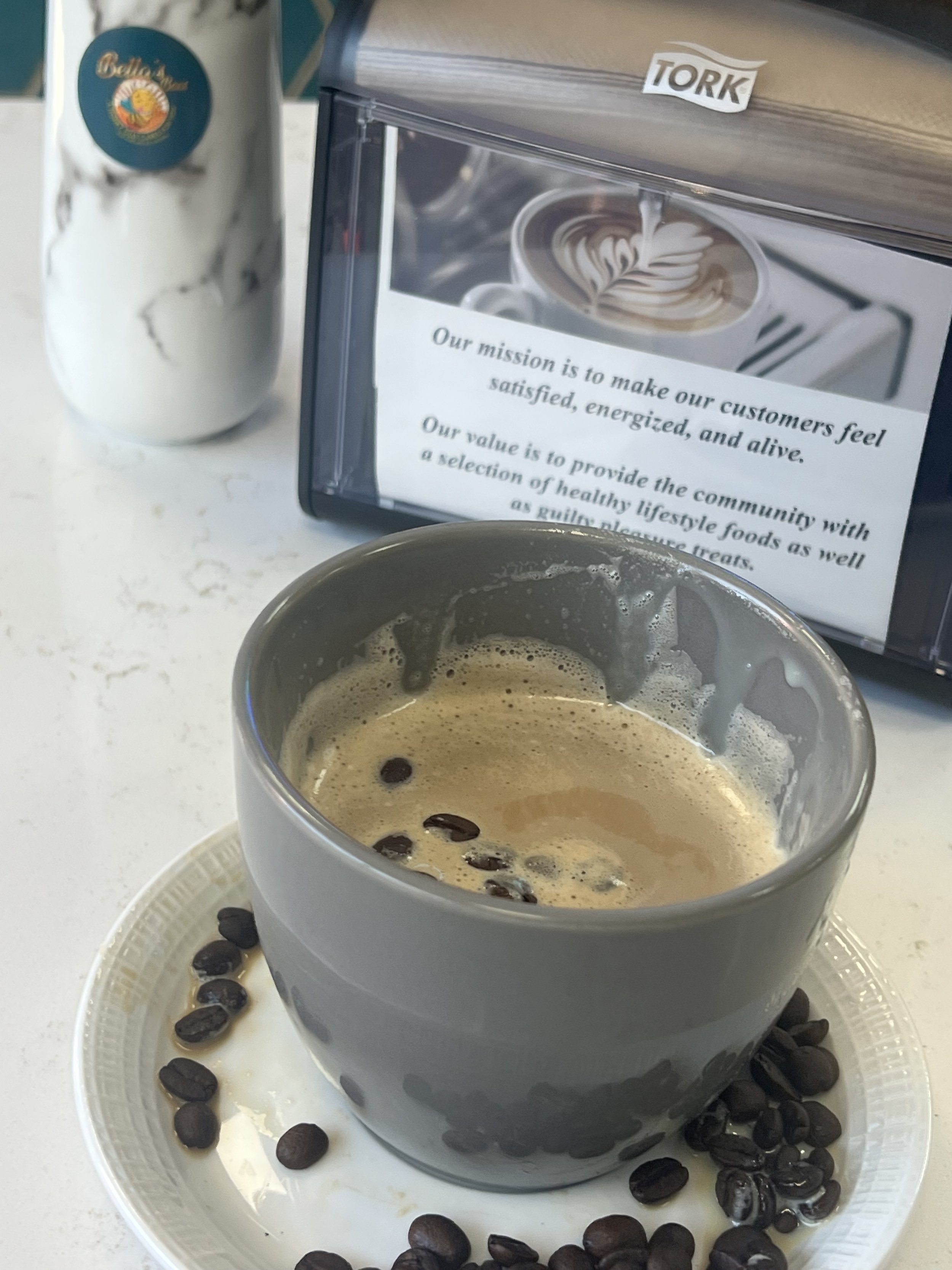 A gray coffee cup filled with espresso on a white saucer surrounded by coffee beans, with a marble-patterned bottle and a napkin dispenser displaying a mission statement in the background.