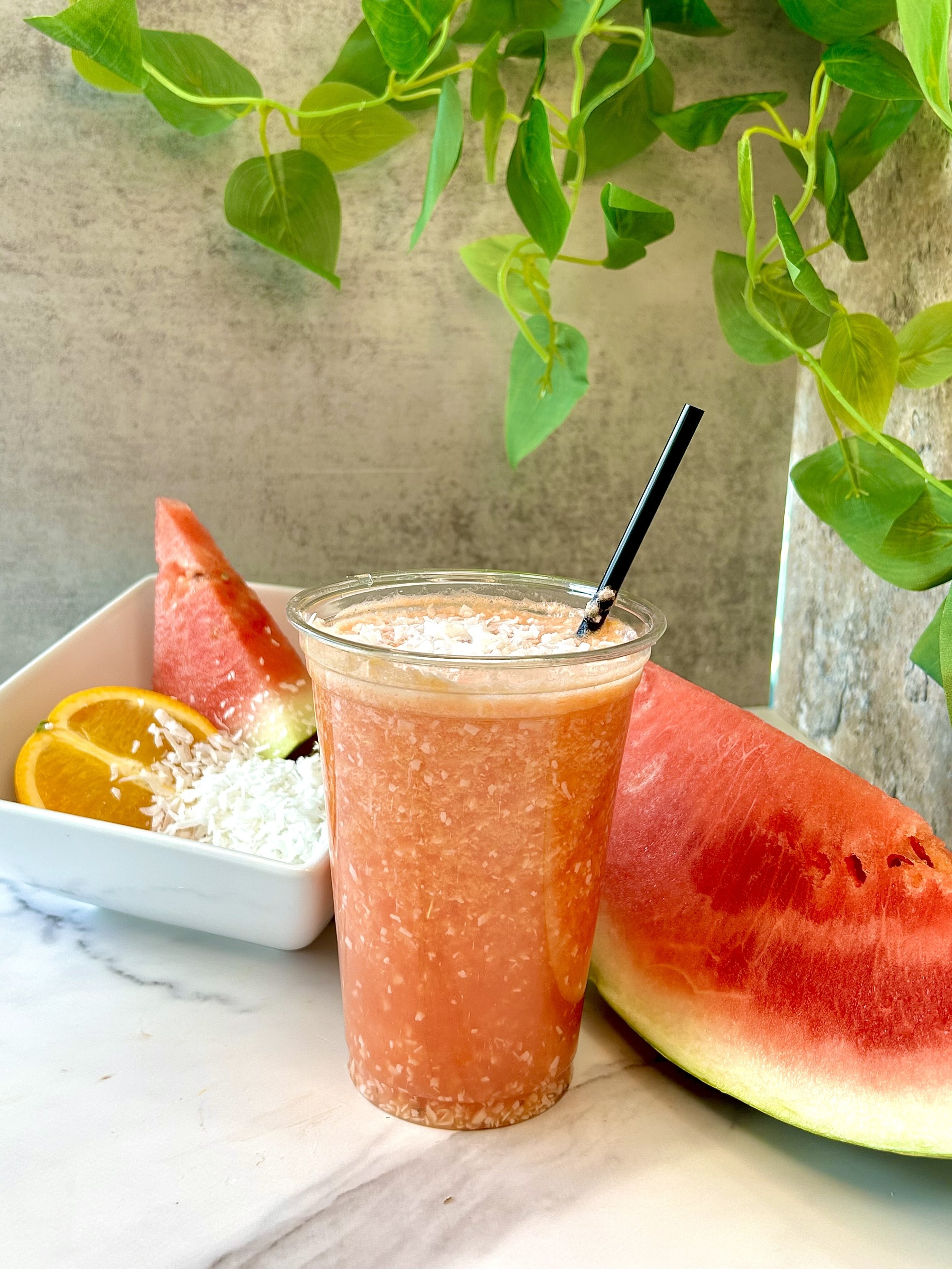 Glass of watermelon smoothie with coconut in a plastic cup with a black straw, surrounded by watermelon slices, orange slices, and shredded coconut on a marble surface; green leaves above.