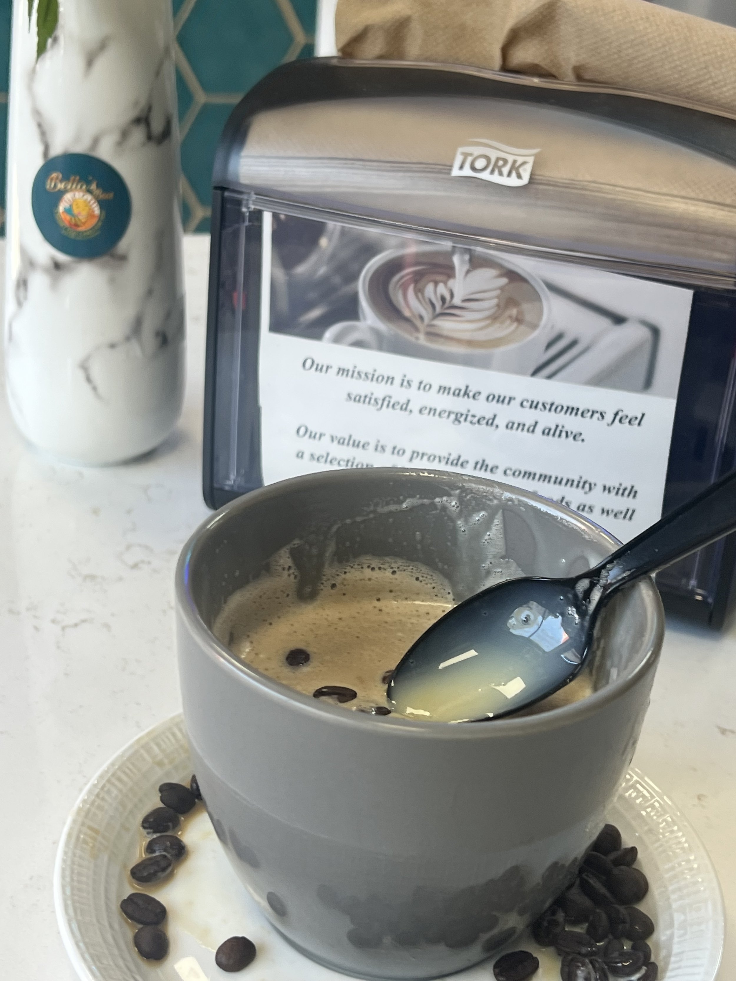A gray mug of coffee with a spoon, coffee beans scattered around, a bottle on the side labeled 'Bella,' and a napkin dispenser with a mission statement text and a coffee image on a countertop.