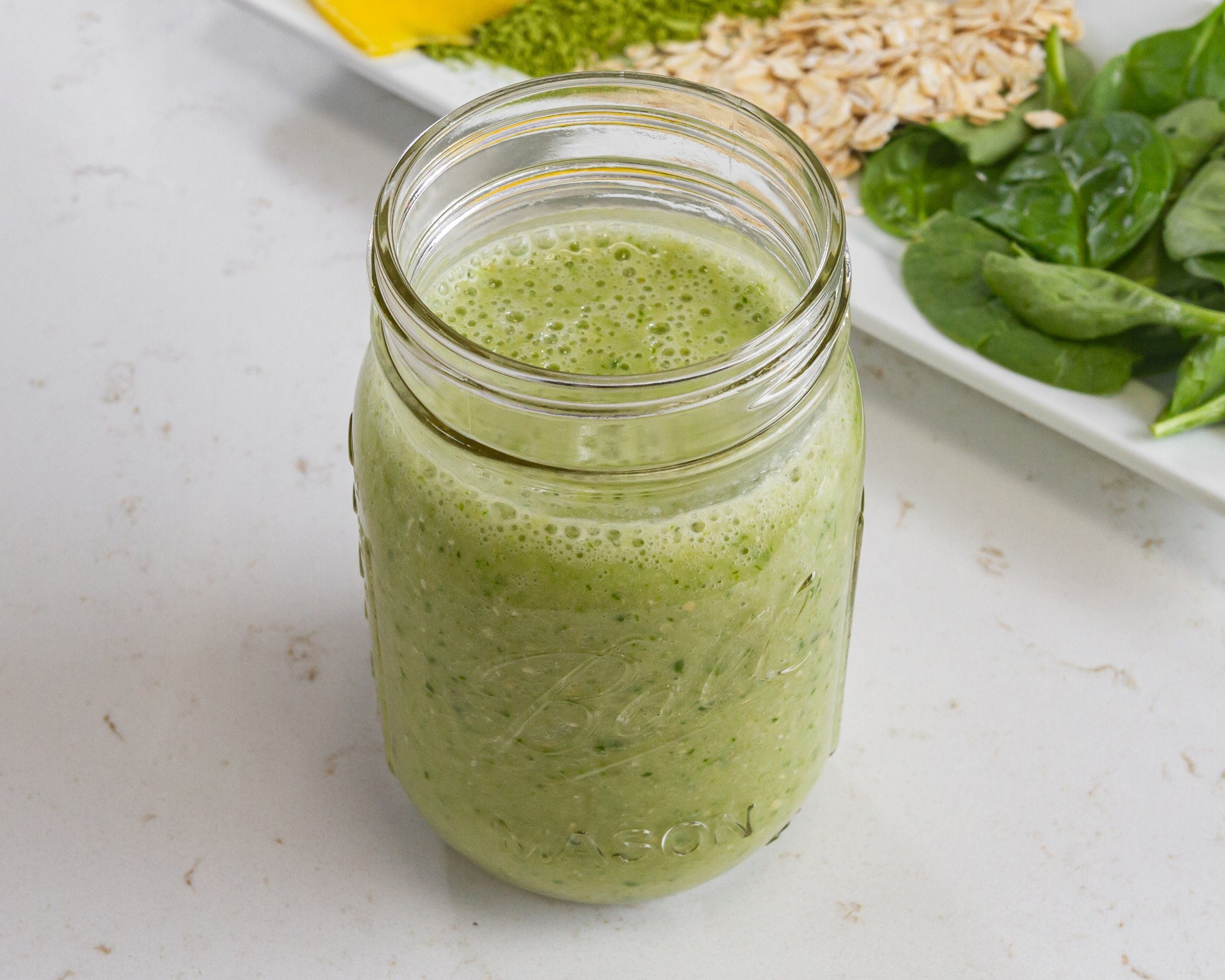 Green smoothie in a mason jar with spinach, oats, and yellow food on a plate.